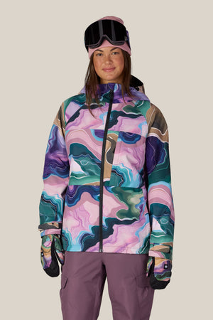 A woman stands facing the camera, smiling slightly, against a plain light background. She is wearing the 686 Women’s Athena™ Insulated Jacket by 686, which features a colorful, abstract pattern, along with purple snow pants, gloves, and goggles.