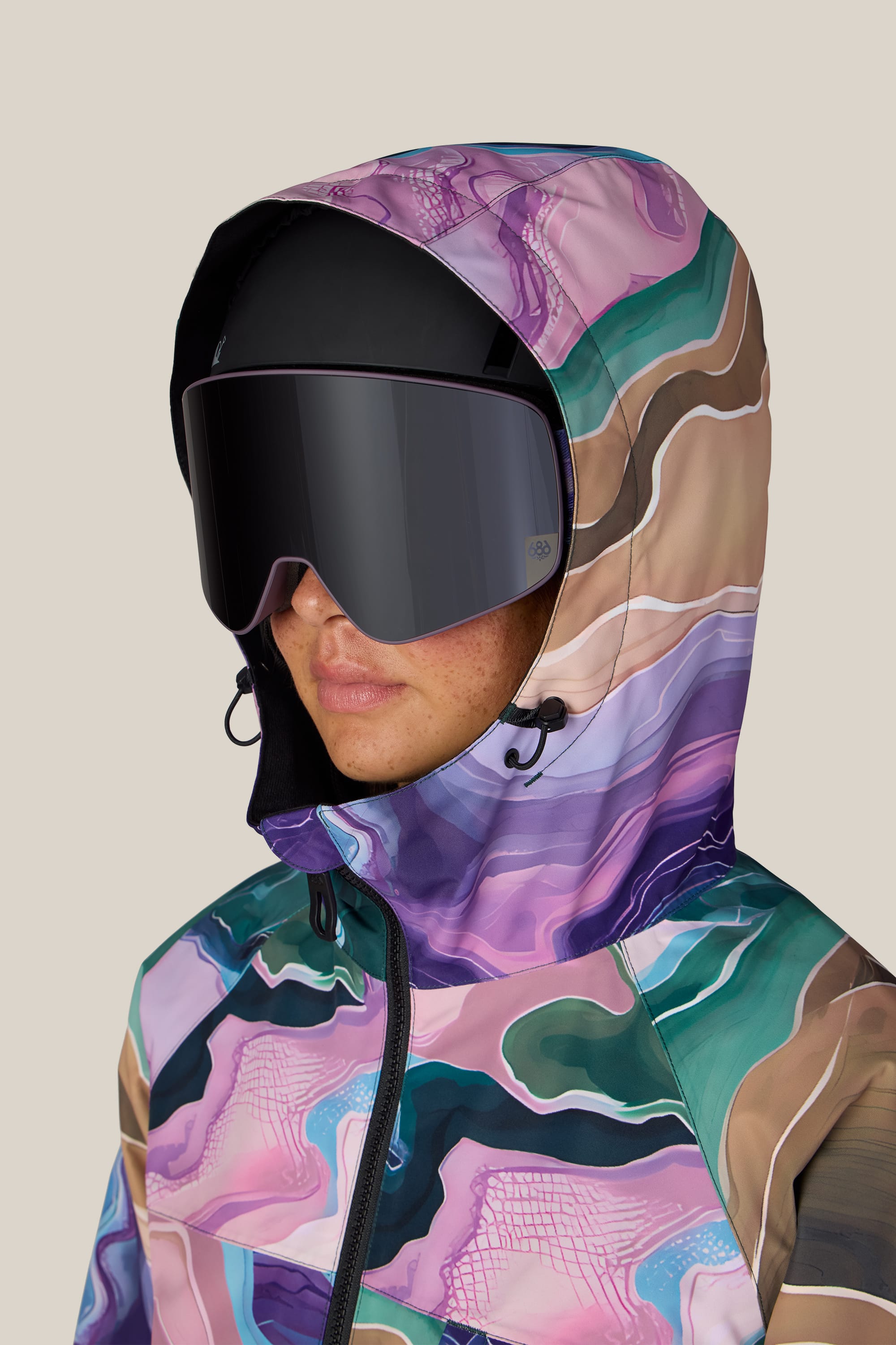 A person wearing the 686 Women’s Athena™ Insulated Jacket, featuring a colorful pattern and hood, paired with large dark ski goggles, is facing left against a plain beige background.