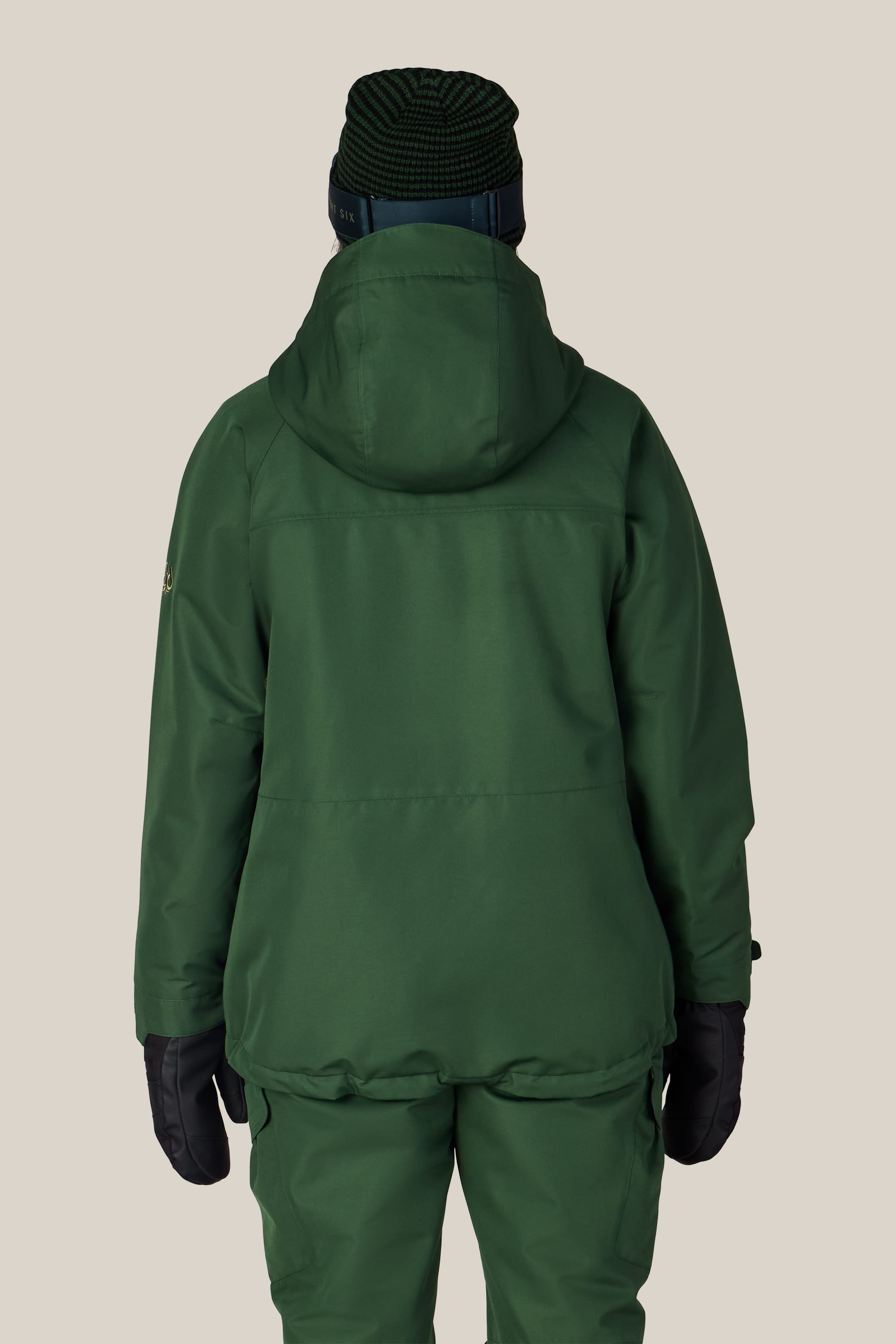 A person is standing with their back to the camera against a plain beige background, wearing the 686 Women’s Athena™ Insulated Jacket by 686, green pants, black gloves, and a dark knit hat.