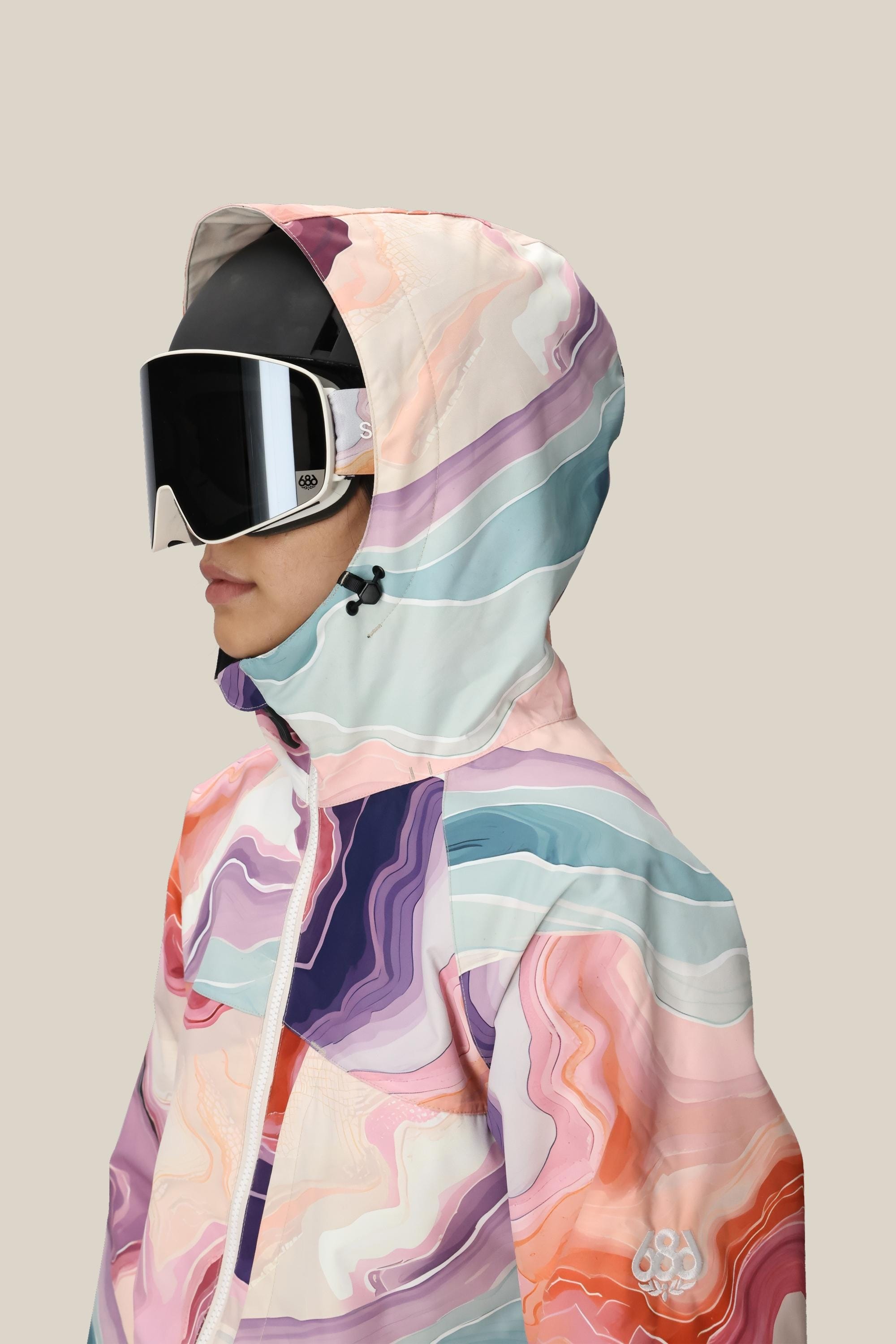 A woman in profile wears the 686 Women's Athena™ Insulated Jacket—an abstract-patterned, waterproof hooded jacket by 686—and large reflective ski goggles, set against a plain beige background.