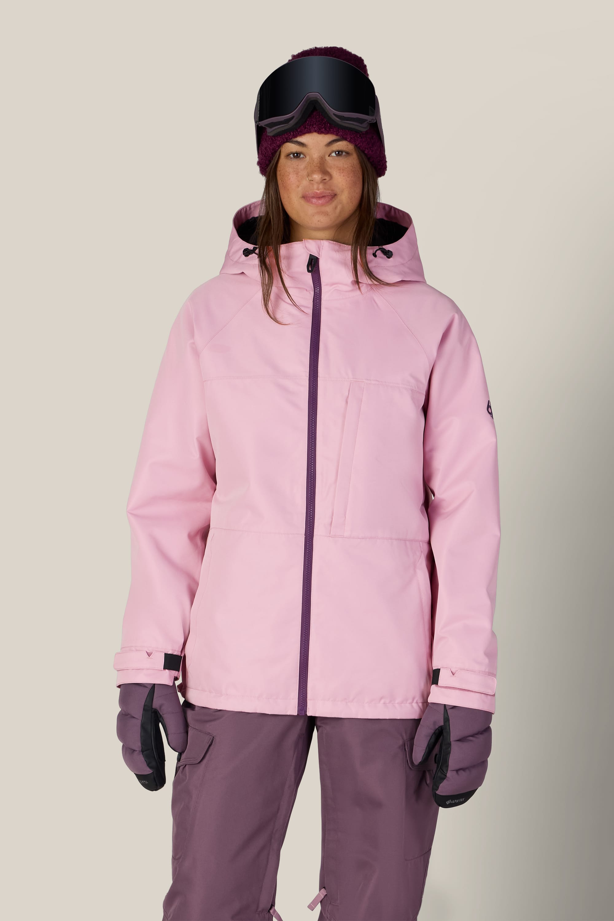 A person wearing the 686 Women’s Athena™ Insulated Jacket by 686, along with purple snow pants, purple gloves, a dark purple beanie, and ski goggles stands facing forward against a plain light background.