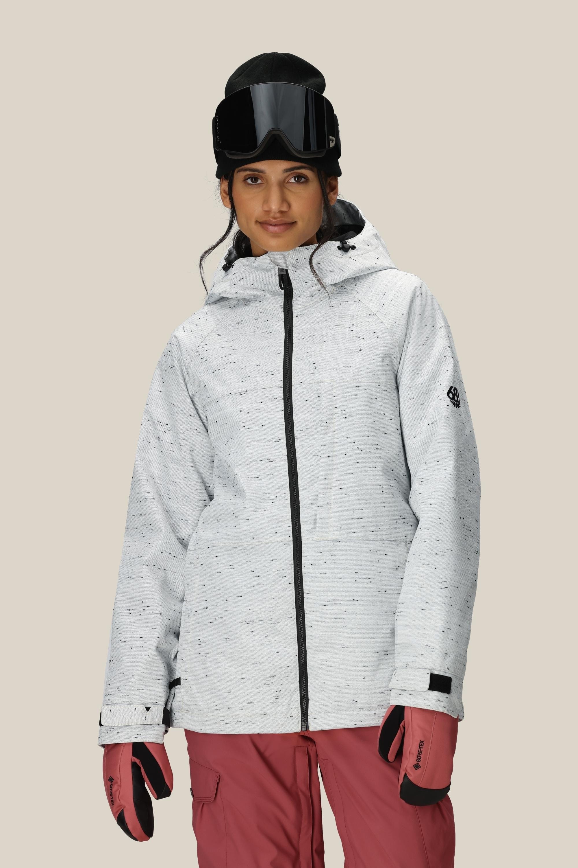 A person wearing the 686 Women’s Athena™ Insulated Jacket in light gray, paired with pink ski pants, pink gloves, and black ski goggles, standing against a plain beige background.