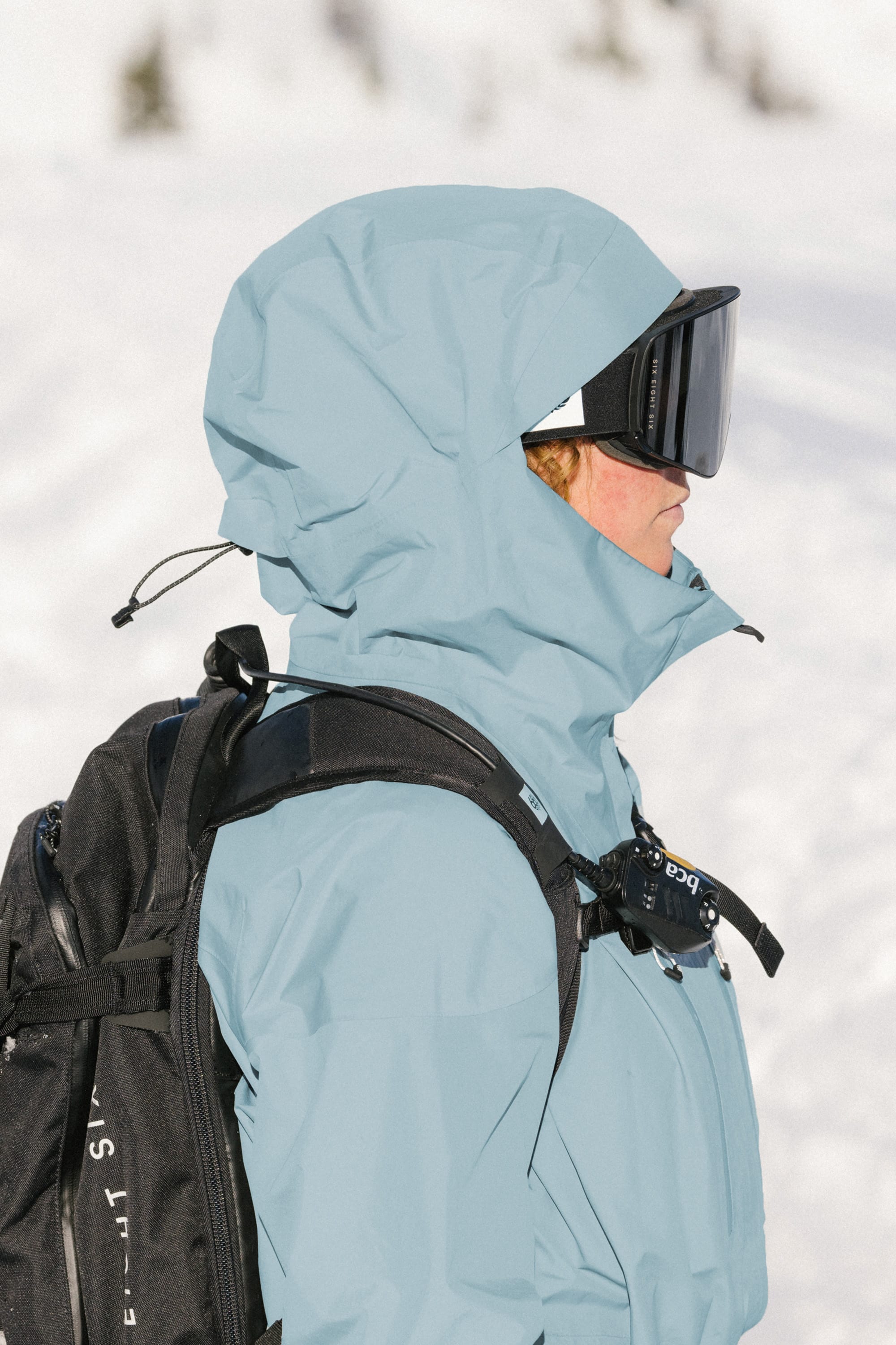 A person wearing a 686 Women's GORE-TEX Skyline Thermagraph® Jacket in light blue, paired with black ski goggles and a black backpack, stands in a snowy outdoor setting.