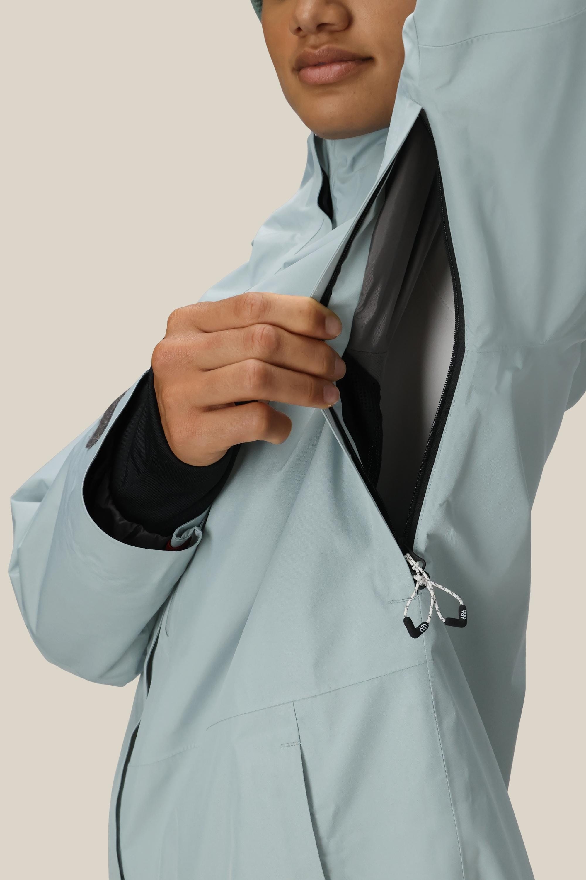 A woman wearing the 686 Women's GORE-TEX Skyline Thermagraph® Jacket in light blue unzips an underarm vent, showing the jacket’s interior against a plain, light background.