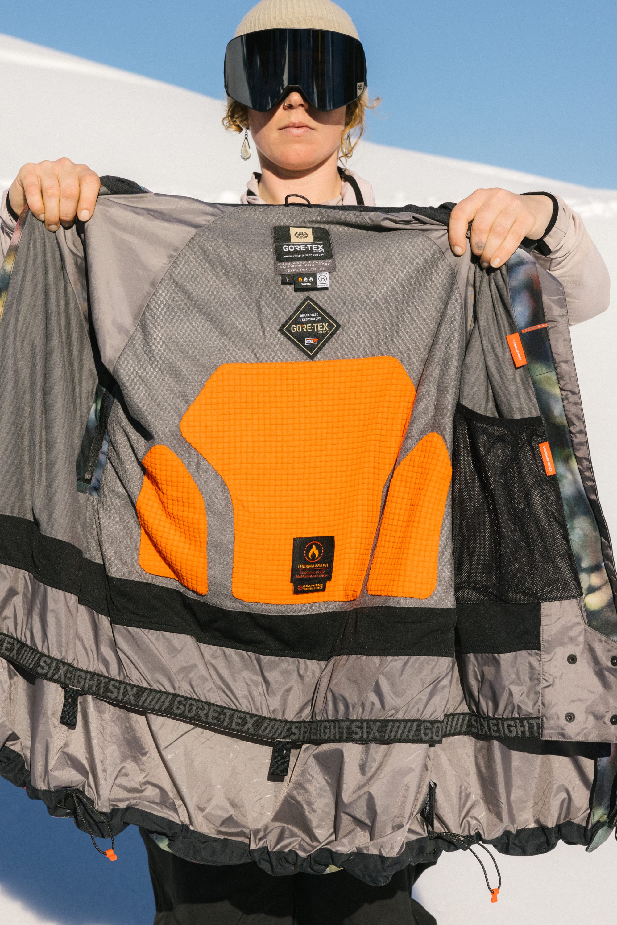 A person in ski goggles and a beige hat displays the orange padded interior and mesh pockets of the 686 Women's GORE-TEX Skyline Thermagraph® Jacket by 686, holding it open against a snowy backdrop.