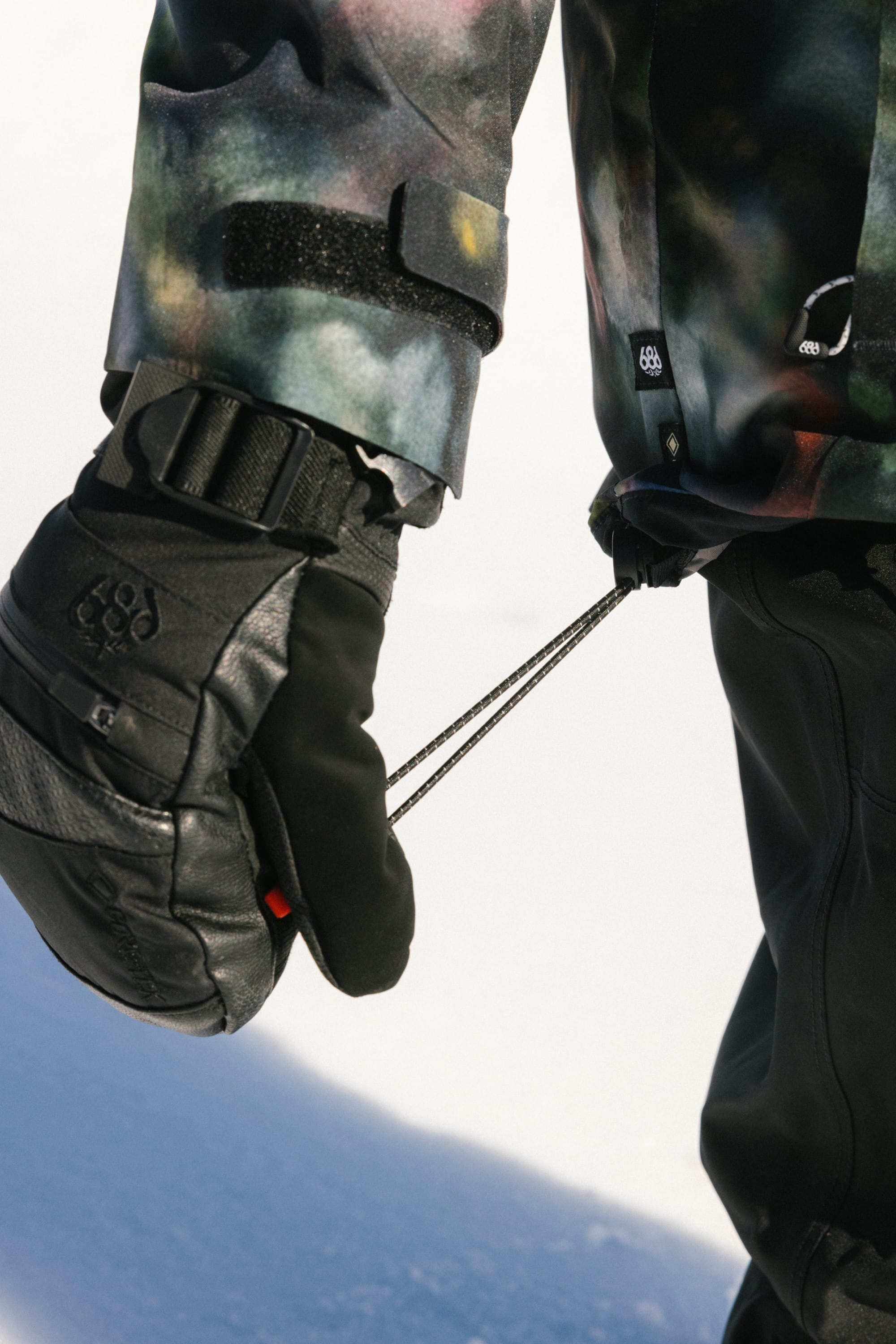 A close-up of someone wearing black winter gloves and the 686 Women's GORE-TEX Skyline Thermagraph® Jacket, adjusting the hem's elastic cord while standing on white snow.