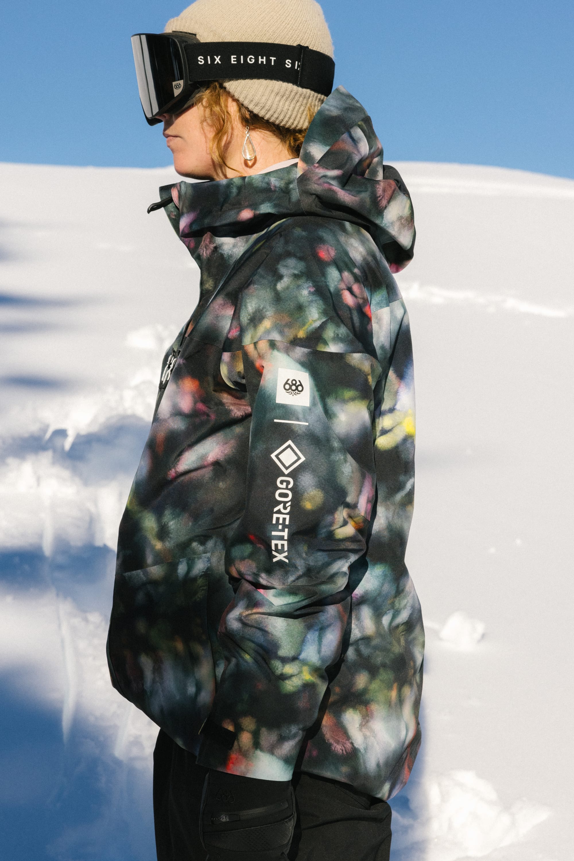 A person wearing the 686 Women's GORE-TEX Skyline Thermagraph® Jacket and ski goggles stands in profile on a snowy landscape under a clear blue sky.