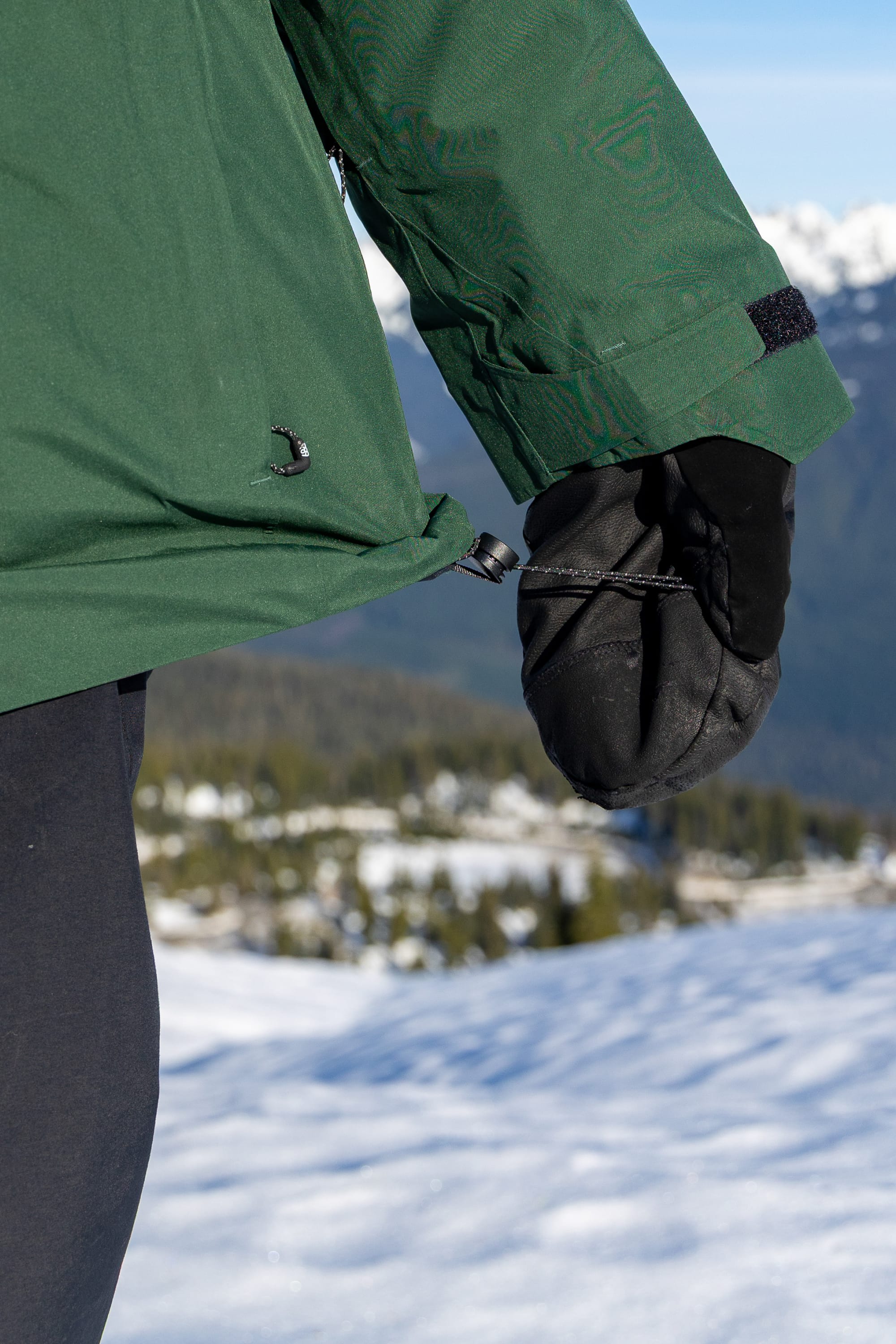 A woman wearing the 686 Women’s GORE-TEX® Skyline Thermagraph® Jacket and black gloves adjusts a drawstring while standing outdoors on a snowy mountain, with trees and distant peaks in the background.