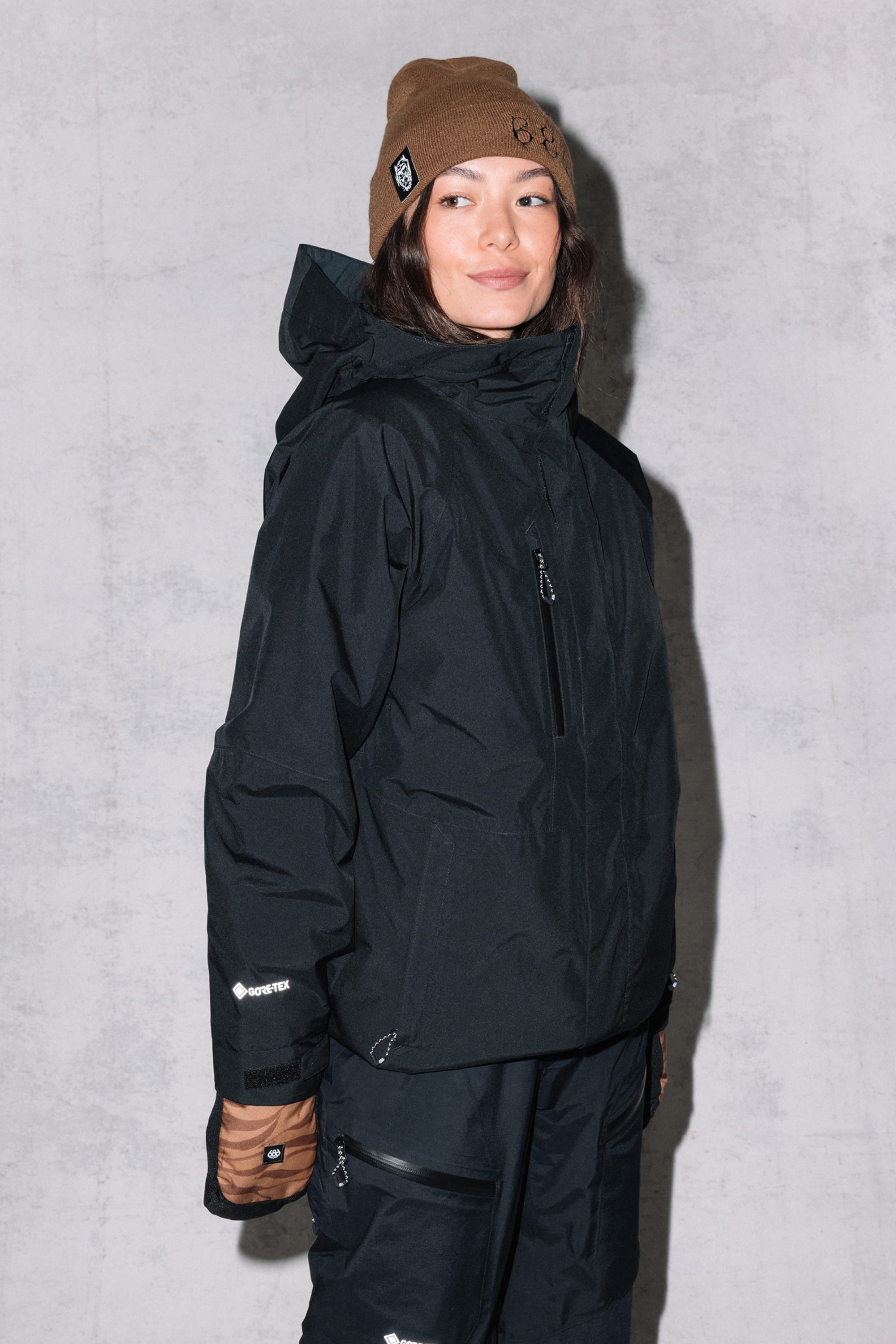 Wearing the 686 Women's GORE-TEX Willow™ Insulated Jacket, black pants, brown gloves, and a brown beanie, a woman stands against a light gray textured wall, looking slightly to the side and smiling.