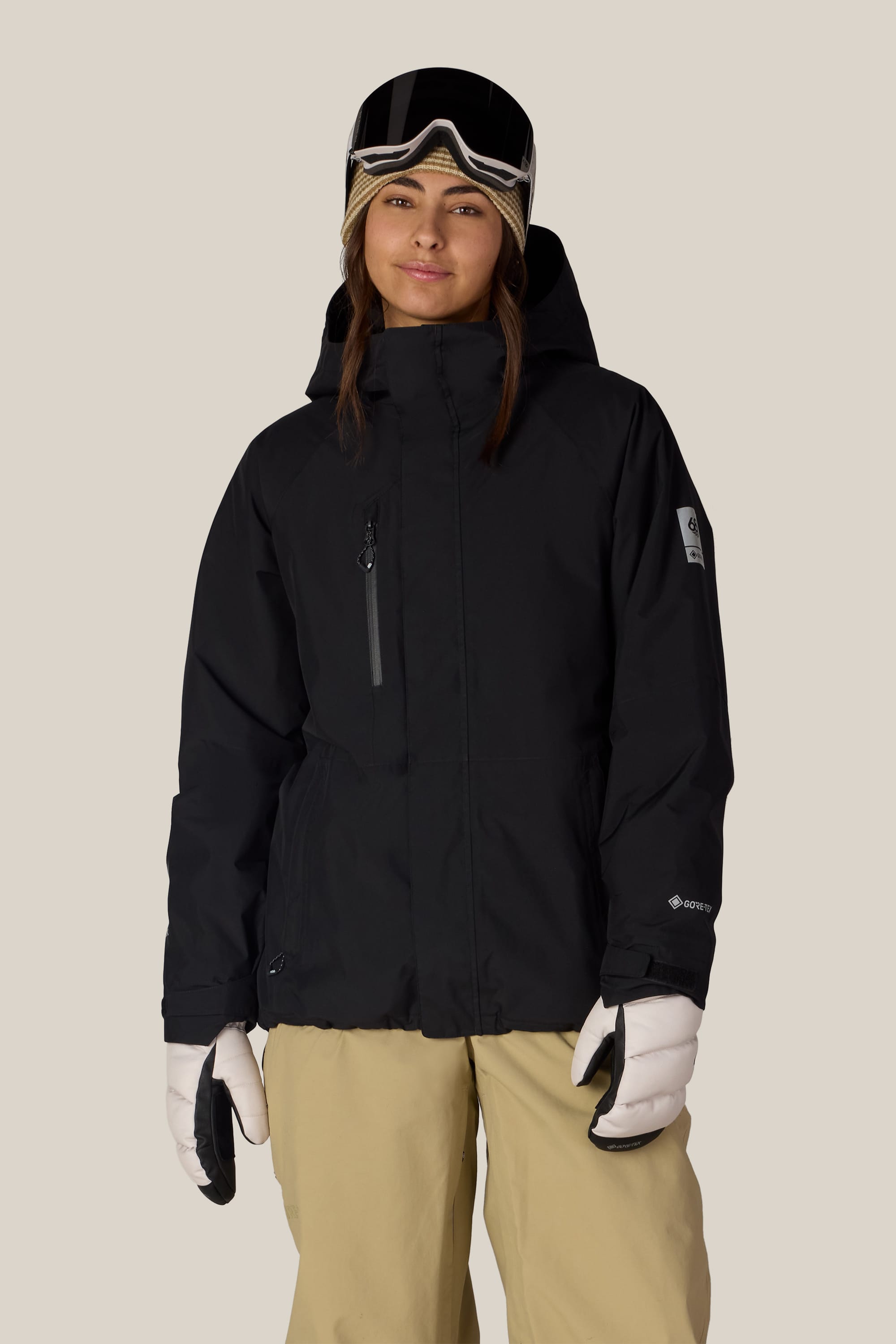 A person wearing the 686 Women’s GORE-TEX® Willow™ Insulated Jacket in black, paired with beige snow pants, white gloves, and ski goggles on their head stands facing forward against a plain light background.