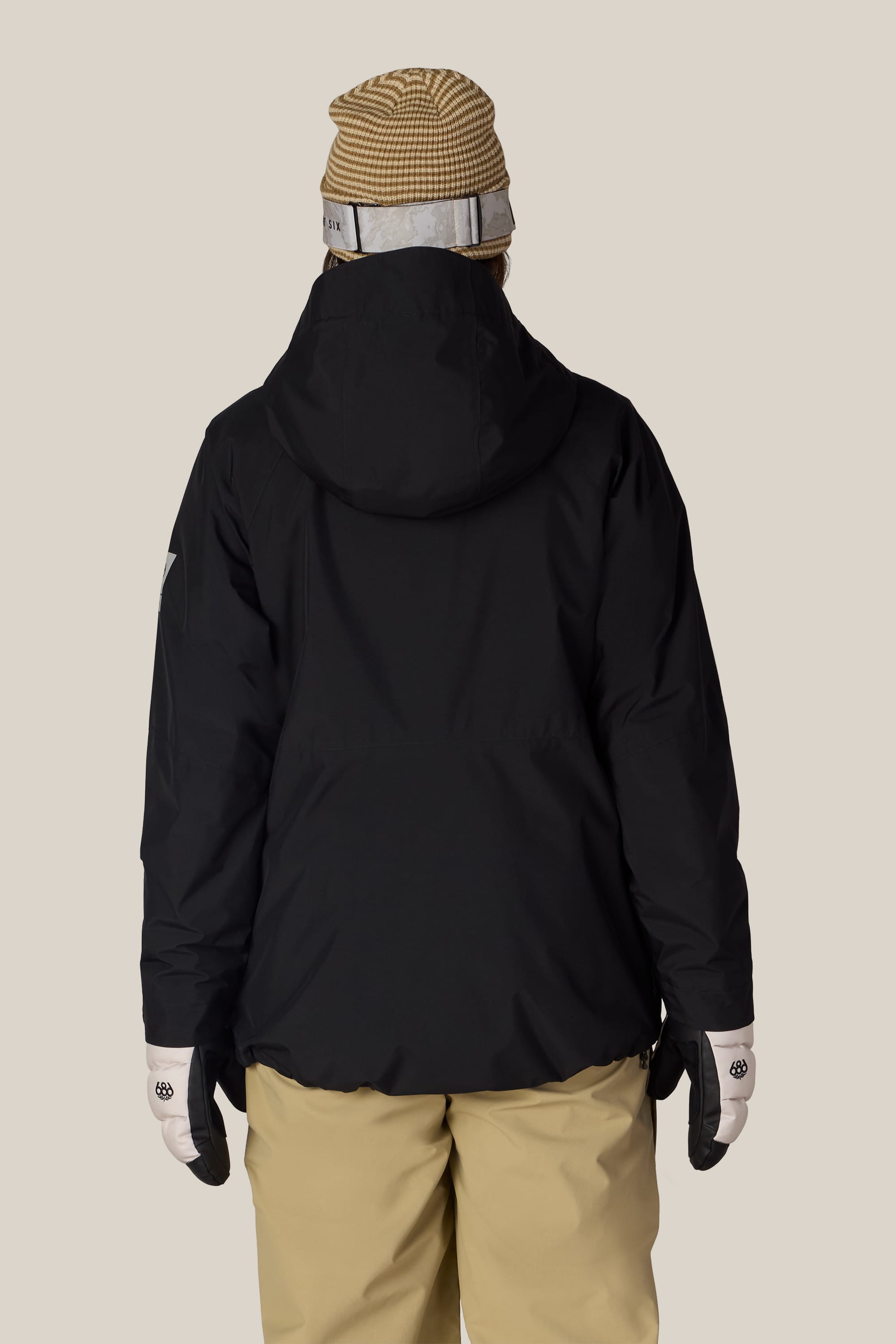 Wearing the 686 Women’s GORE-TEX® Willow™ Insulated Jacket in black, a person stands facing away from the camera against a plain light background, styled with beige pants, white gloves, and a tan striped beanie.