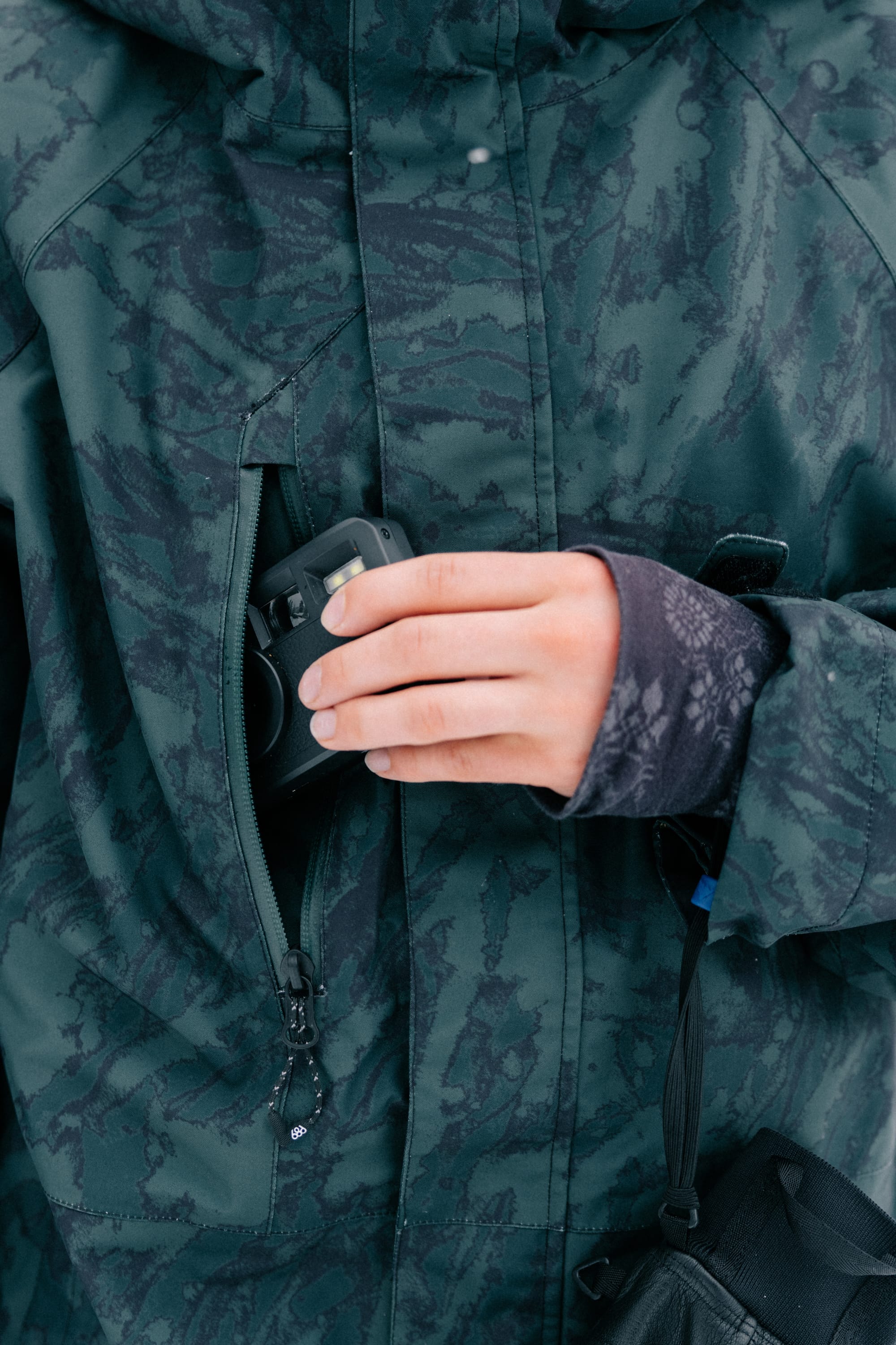 Wearing the 686 Women’s GORE-TEX® Willow™ Insulated Jacket, a person slips a small black camera into the jacket’s zippered chest pocket, with only their hand and part of the patterned outerwear visible.