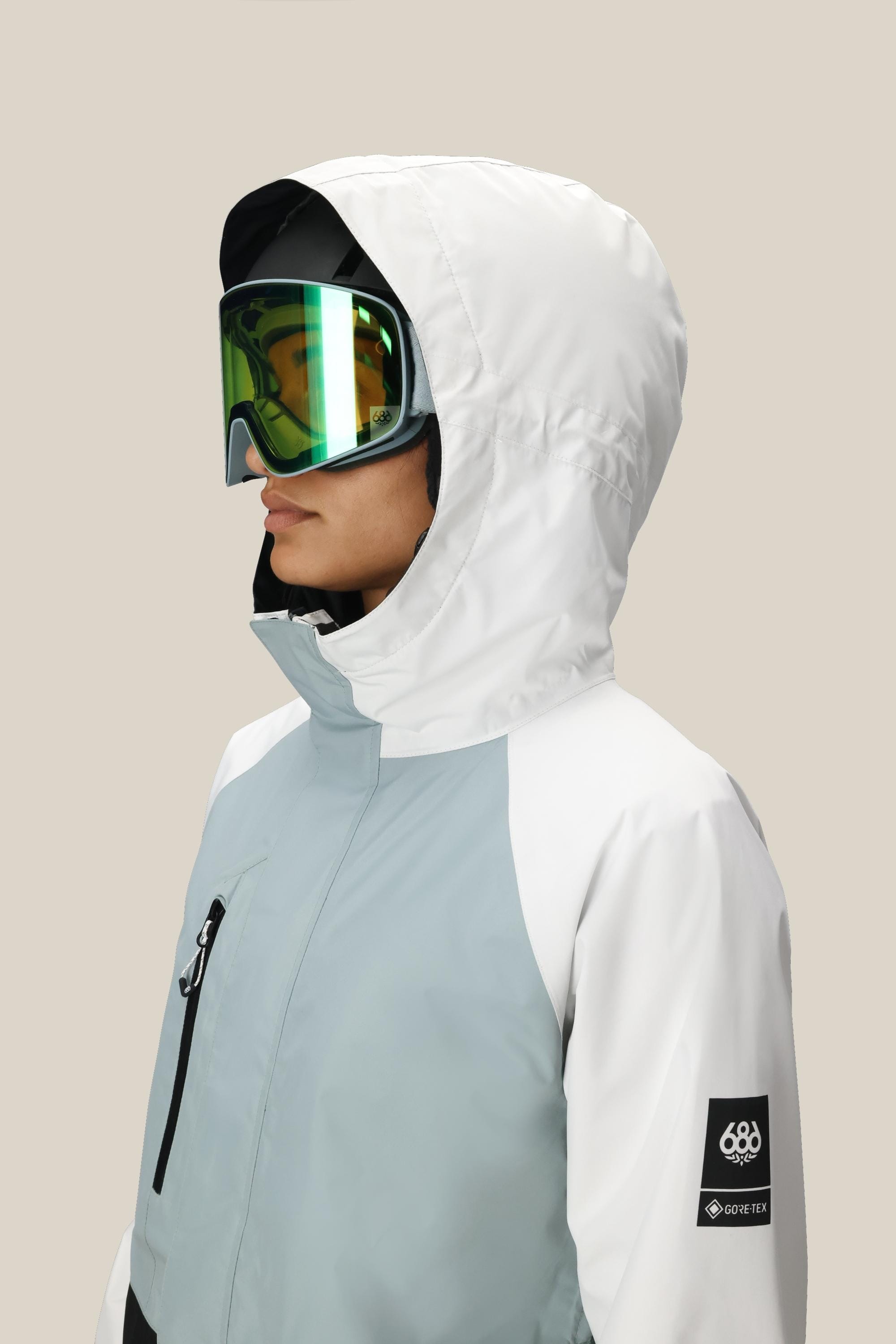 A woman in profile against a beige background wears the 686 Women's GORE-TEX Willow™ Insulated Jacket in white/light gray with reflective ski goggles. The sleeve displays GORE-TEX and 686 logo patches.
