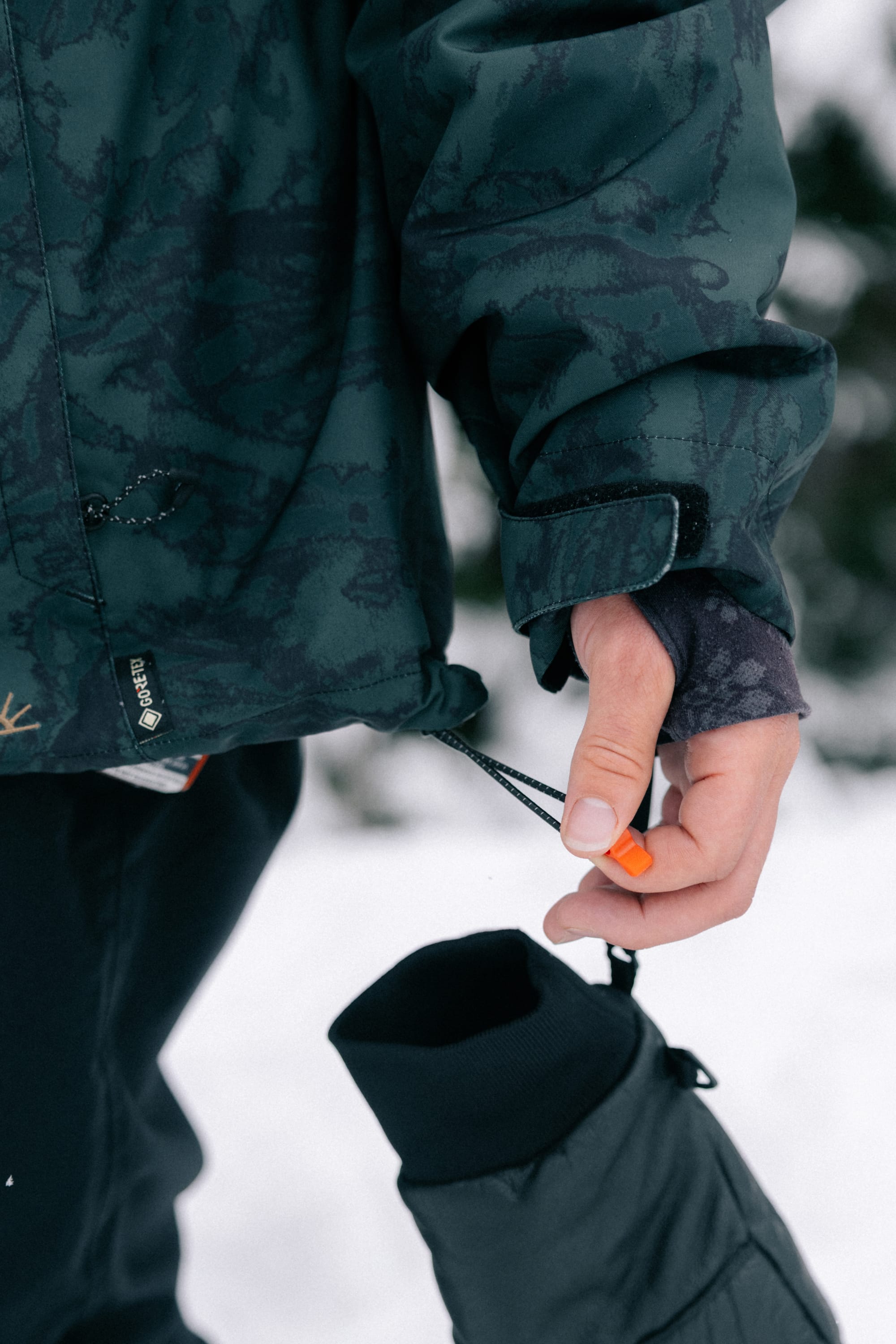 Wearing the 686 Women’s GORE-TEX® Willow™ Insulated Jacket in a dark green pattern, a person tightens the sleeve drawstring while holding a black mitten, standing outdoors in the snow.