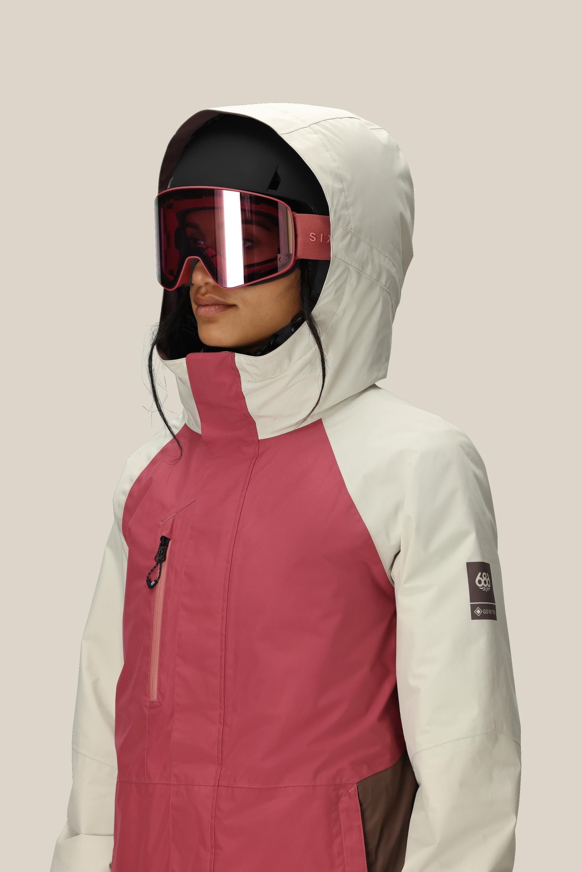 A woman in the 686 Women's GORE-TEX Willow™ Insulated Jacket, wearing a black ski helmet and large red ski goggles, stands against a plain light background.