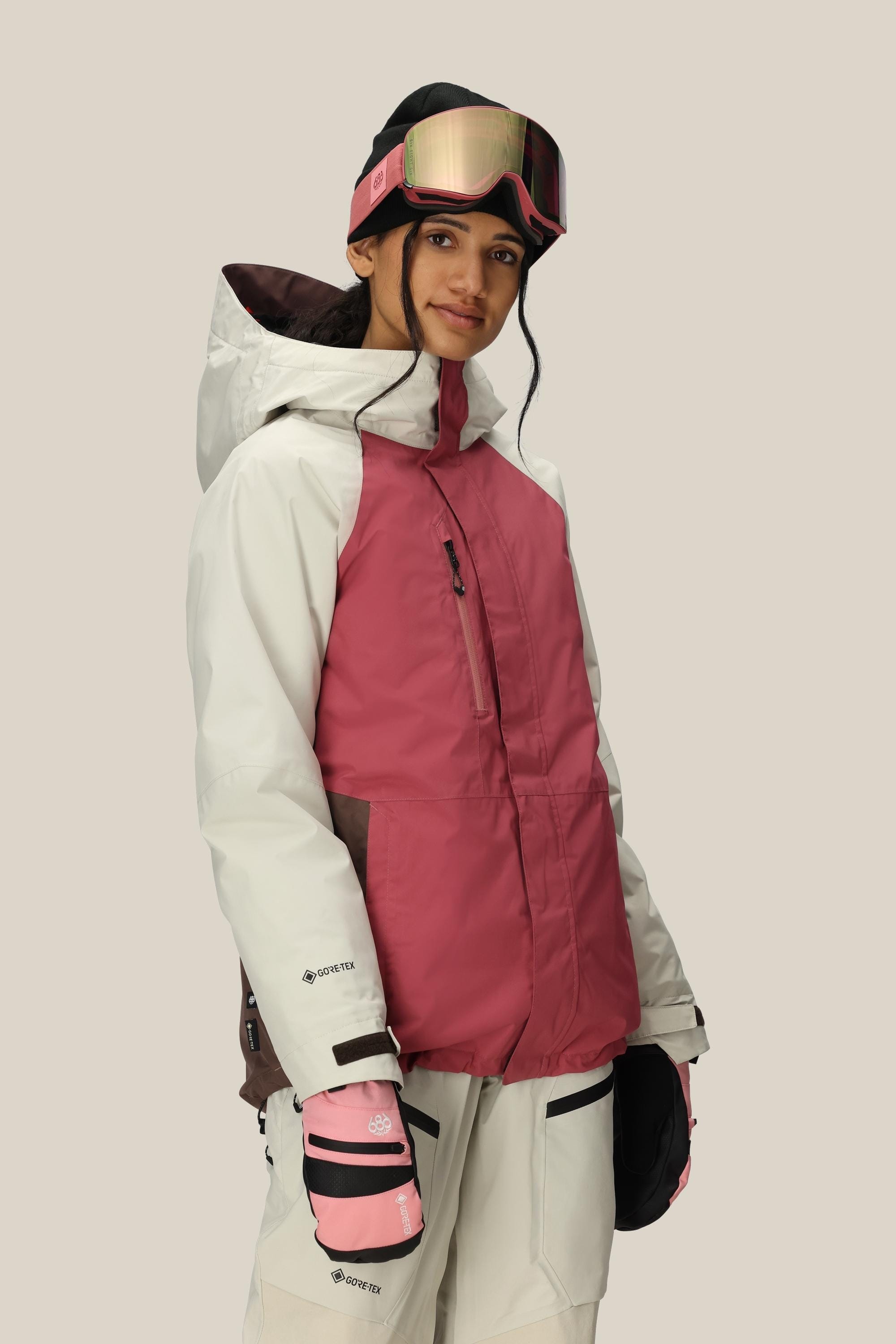 A woman in a 686 Women's GORE-TEX Willow™ Insulated Jacket, white ski pants, pink and white gloves, and pink ski goggles stands against a light background, smiling slightly.
