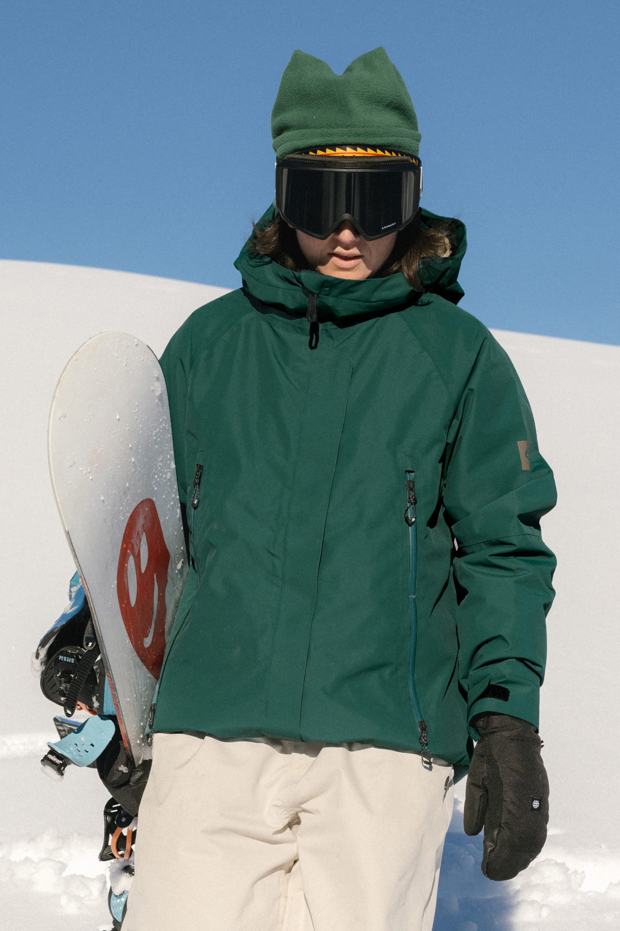 Wearing the 686 Women's Whisper™ Insulated Jacket and a green hat, a person holds a snowboard on snow. They sport large black goggles and brown gloves, with a clear blue sky and snowy slope behind them.