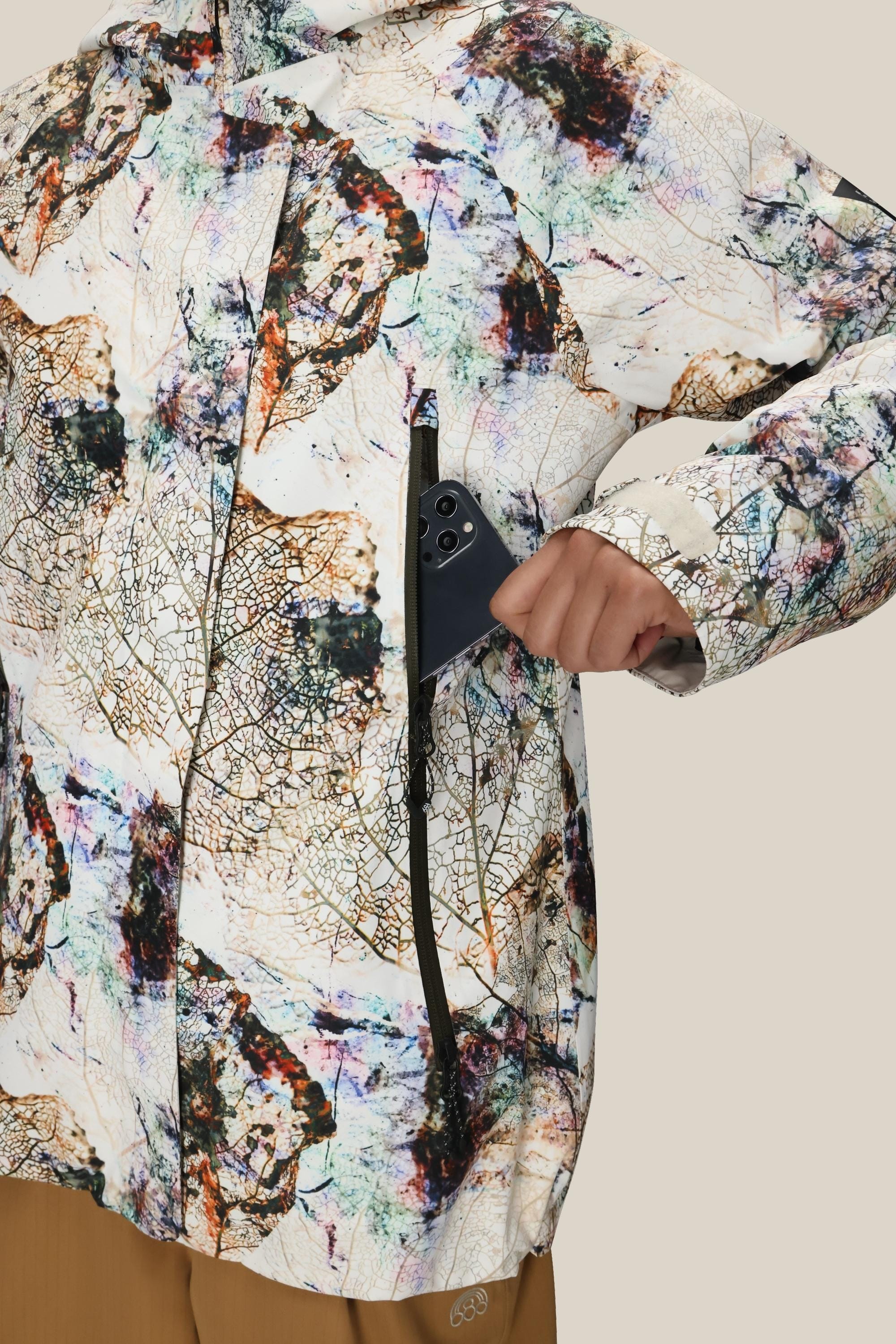 A woman in tan pants slides a black smartphone into the zippered chest pocket of a colorful, abstract-patterned 686 Women's Whisper™ Insulated Jacket by 686, against a plain neutral background.