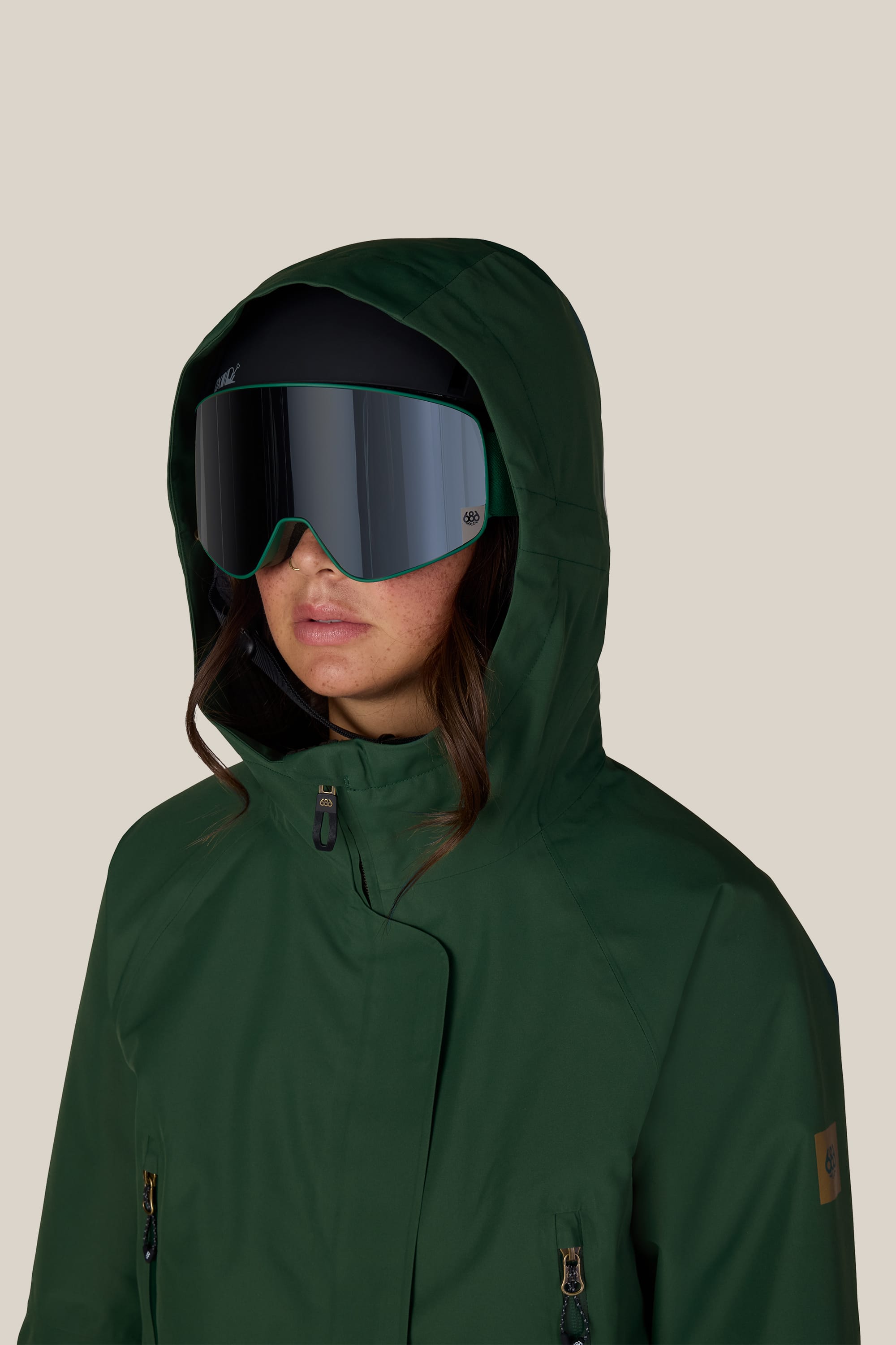 A woman wearing the 686 Women’s Whisper™ Insulated Jacket in dark green and large reflective ski goggles, facing forward against a plain beige background.