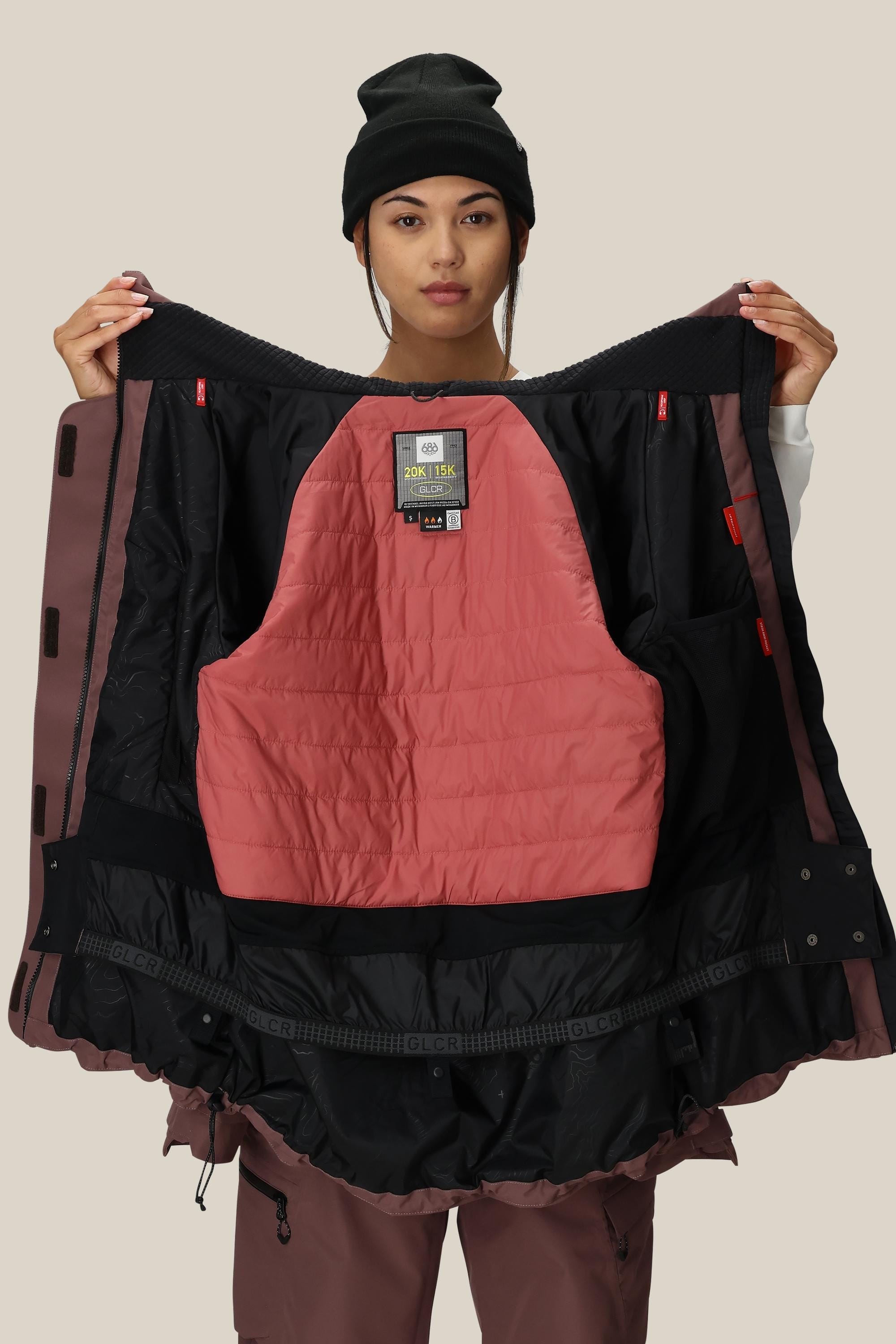 A woman in a black beanie and mauve snow pants models the 686 Women's Whisper™ Insulated Jacket, showing its quilted lining and multiple pockets, facing the camera against a plain beige background.