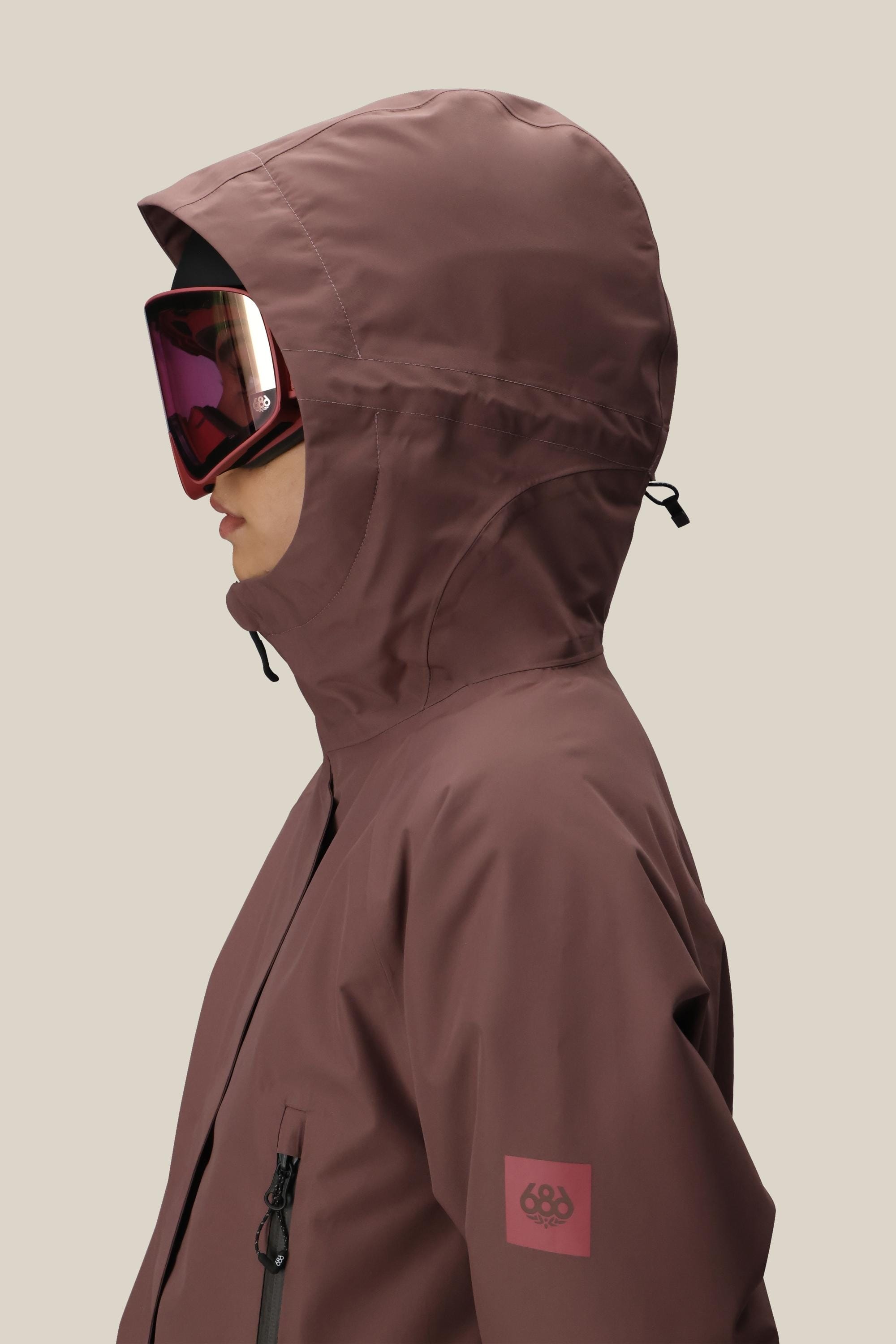 A woman in profile wears the 686 Women's Whisper™ Insulated Jacket and large reflective ski goggles, standing against a plain beige background.