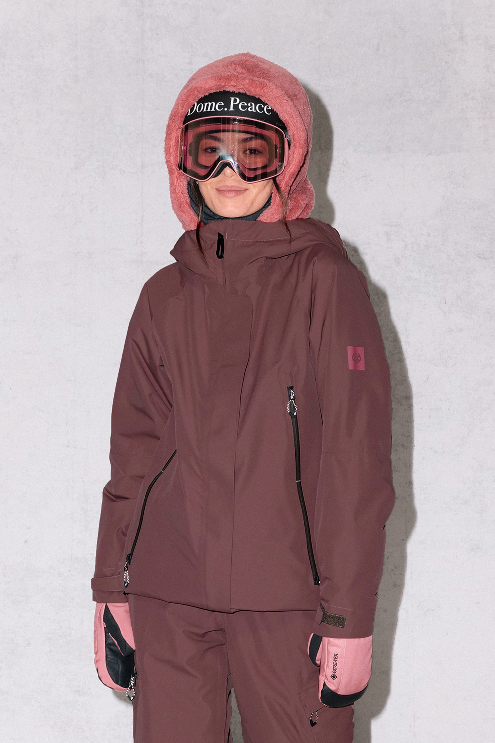 A person stands against a light gray background, smiling slightly in the 686 Women's Whisper™ Insulated Jacket and pants, pink gloves, a fuzzy pink hood, and ski goggles with "Dome. Peace" on the strap.