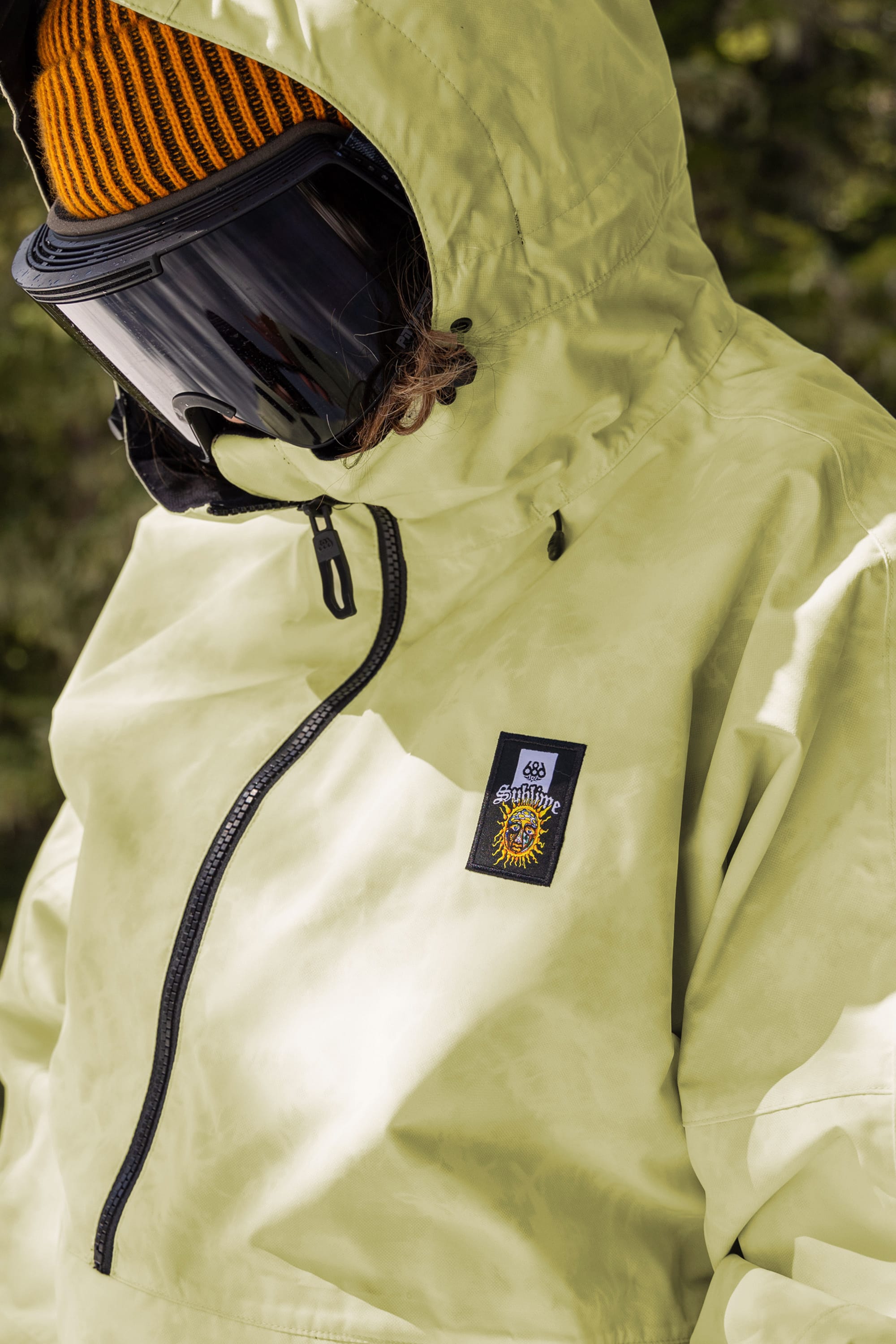 A person wearing the 686 Womens Sublime Shell Anorak in light green, black ski goggles, and an orange knit hat stands outdoors. The jacket features a patch with a sun and Sunshine. Blurred trees are in the background.