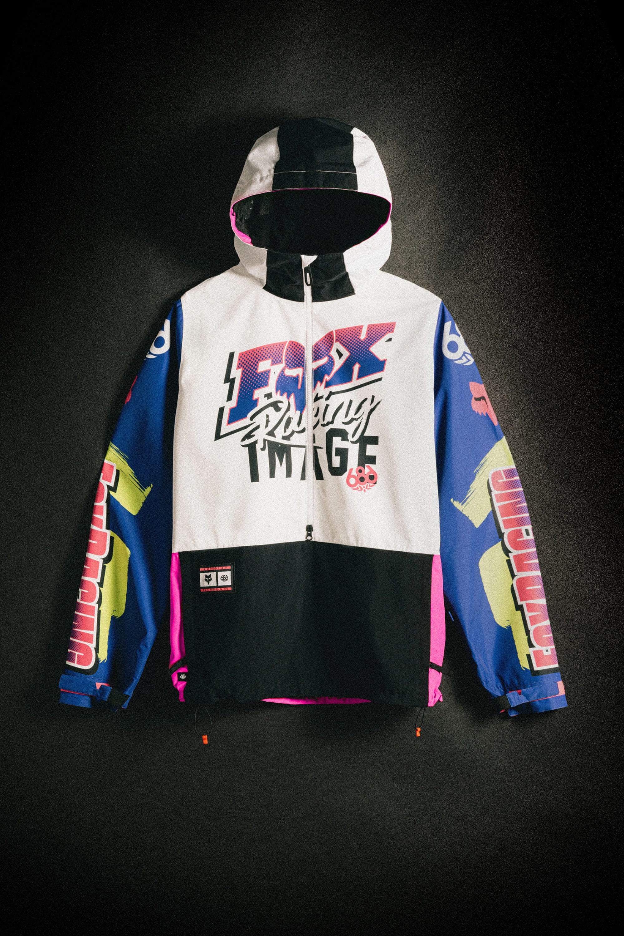 The 686 Men's Fox Racing Shell Anorak features bold "FOX Racing Image" graphics on the front, vibrant blue, pink, yellow, and white panels, and racing designs on the sleeves. The 686 jacket is displayed flat against a dark background.