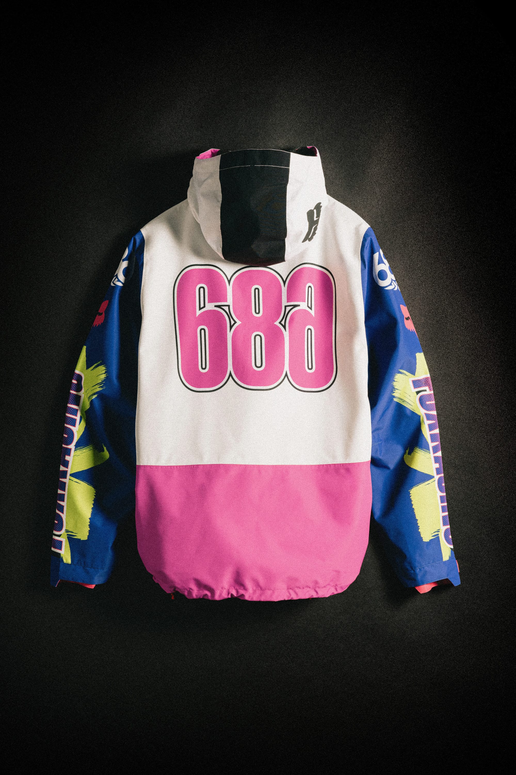 The 686 Men's Fox Racing Shell Anorak by 686 features a large pink "686" on the back, a white upper section, blue sleeves with yellow graphics, and a pink lower section, shown against a dark background.