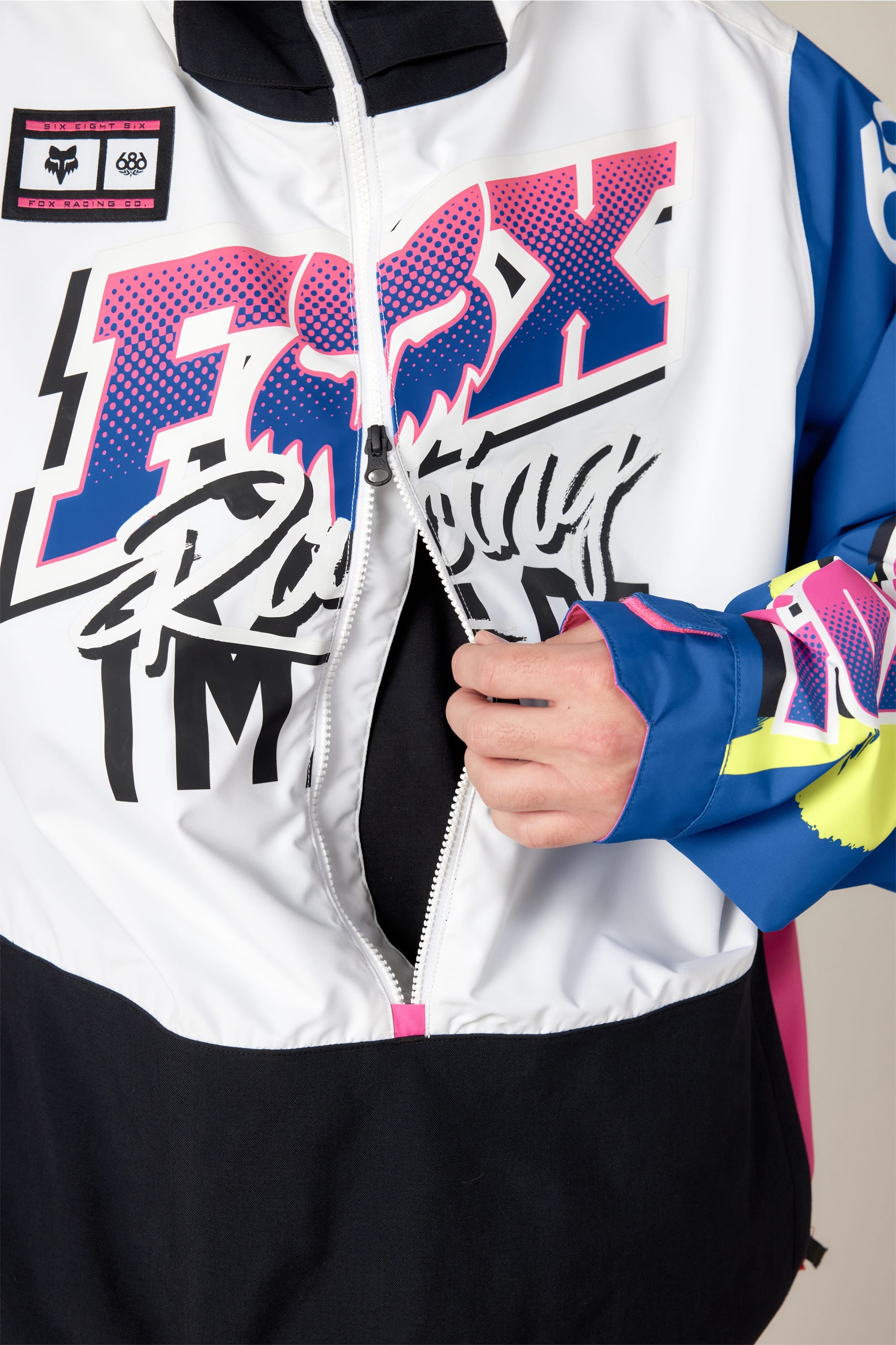 A person unzipping a vibrant 686 Men's Fox Racing Shell Anorak, showcasing bold pink, blue, black, and yellow graphics, with their right hand on the zipper.