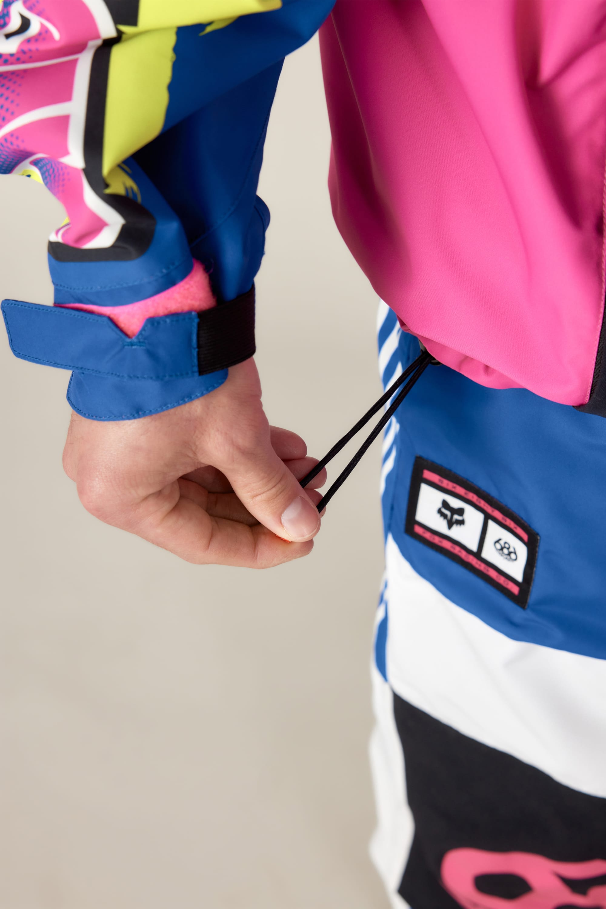 A person pulls the elastic drawstring on a 686 Men's Fox Racing Shell Anorak, featuring vibrant blue, pink, yellow, and white patterns, with a branded patch near their hand.