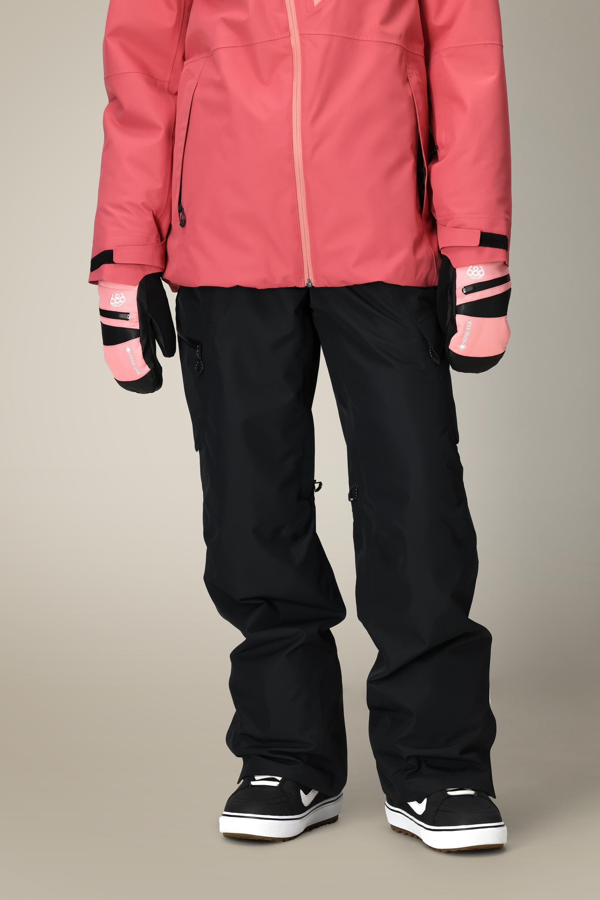 A person wearing a pink jacket, pink gloves, 686 Women's Geode Thermagraph® Pant from 686 in black, and black and white snow boots stands against a plain background with their upper body partially out of frame.