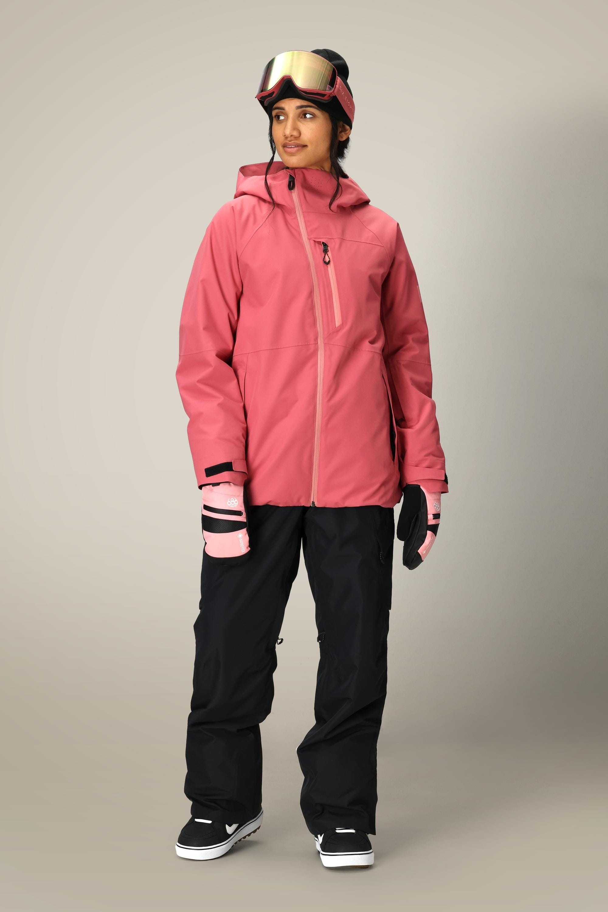 A person wearing a pink hooded ski jacket, 686 Women's Geode Thermagraph® Pant by 686, pink and black gloves, black boots, and ski goggles stands against a plain background.