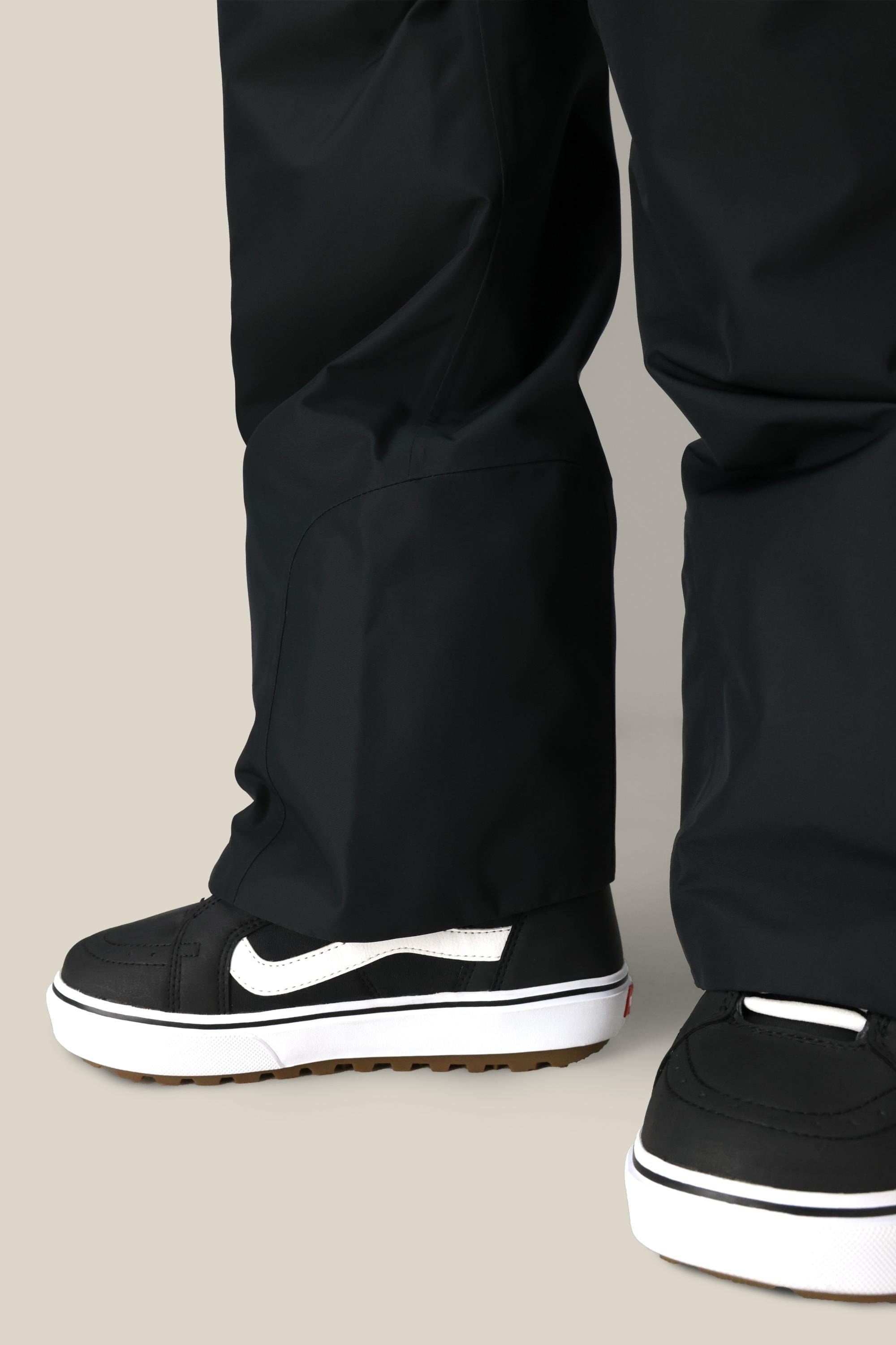 A close-up of a person wearing the 686 Women's Geode Thermagraph® Pant by 686, paired with black skate-style sneakers featuring white soles and a white swoosh logo, standing on a light-colored surface.