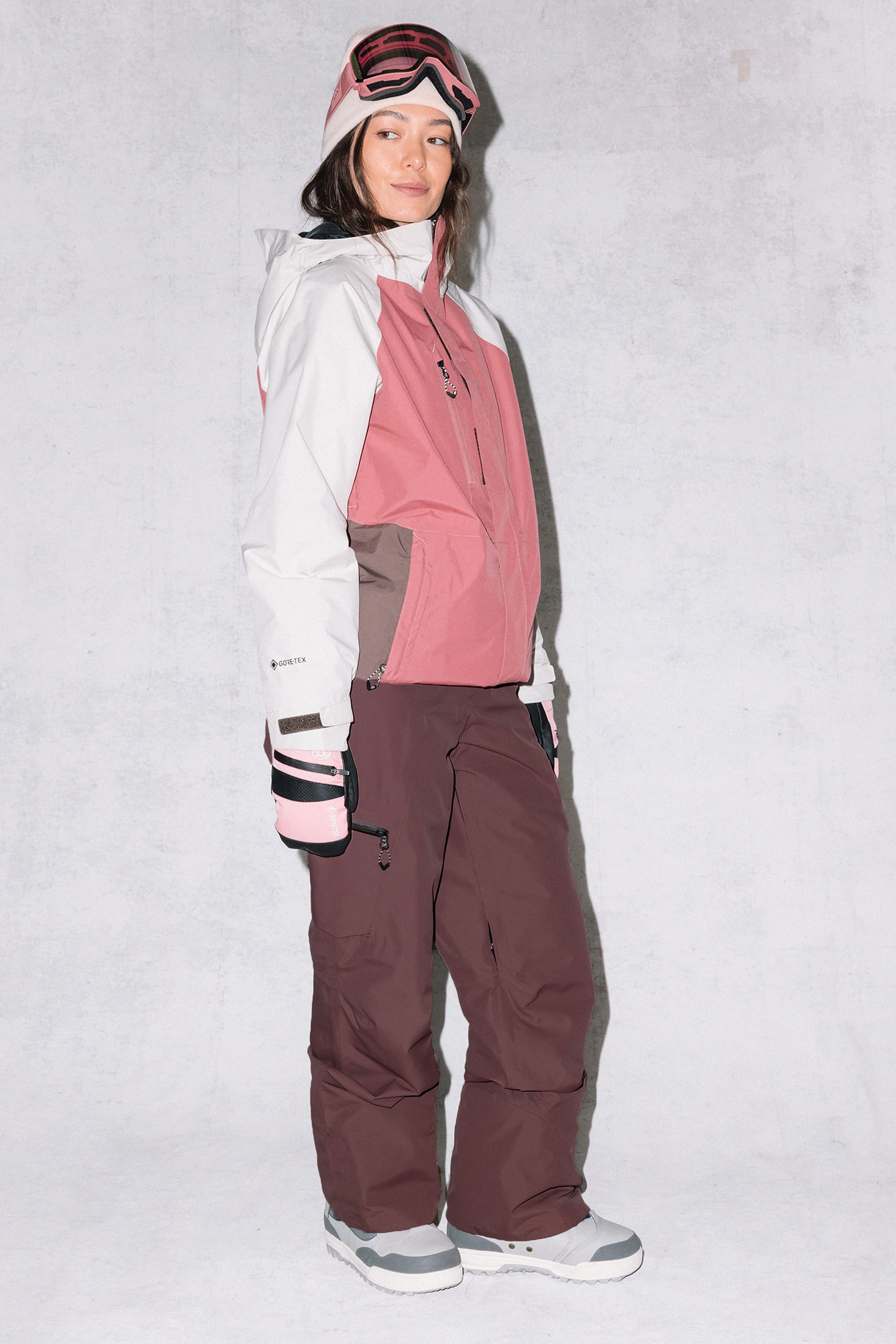 A woman in a pink and white ski jacket, 686 Women's Geode Thermagraph® Pant by 686, and gray boots stands against a light background. Ski goggles rest on her head as she looks to the side with a relaxed expression.
