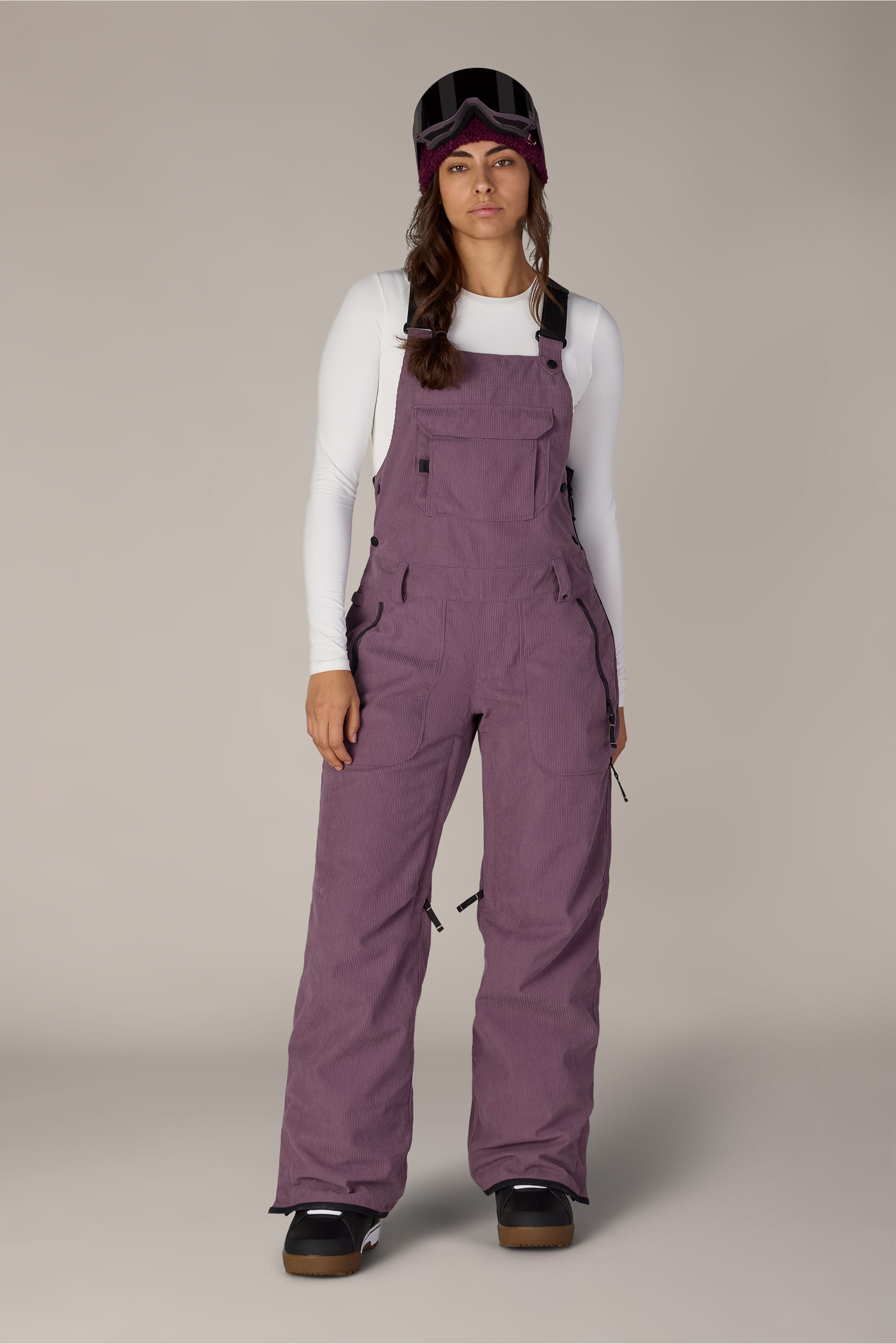 A woman in a white long-sleeve shirt, 686 Women’s Black Magic™ Insulated Bib by 686, black snow boots, and a ski mask with goggles stands against a plain background.