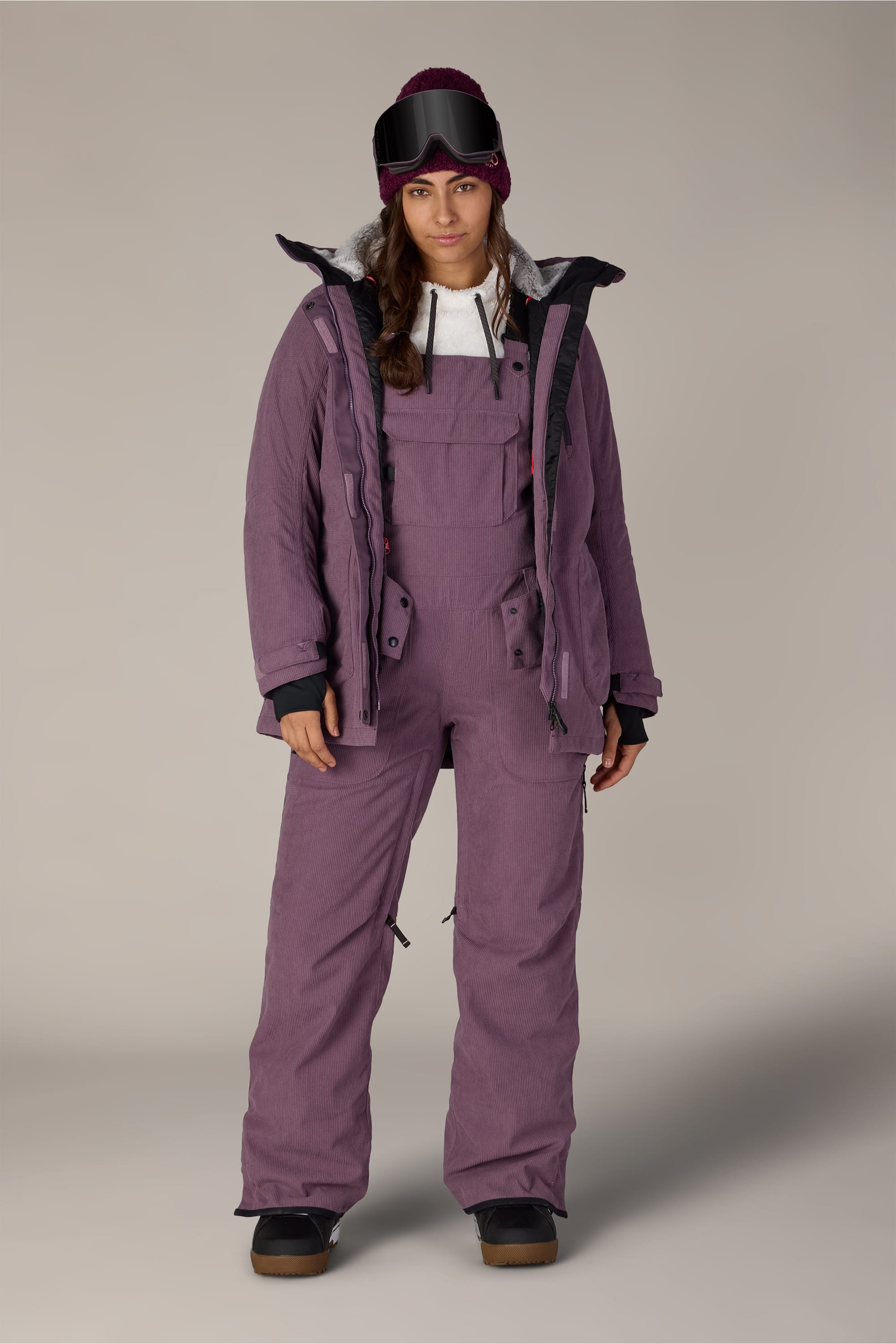 A person stands facing forward against a plain background, wearing the 686 Women’s Black Magic™ Insulated Bib by 686 along with a matching ski jacket, knit hat, ski goggles, and snow boots.