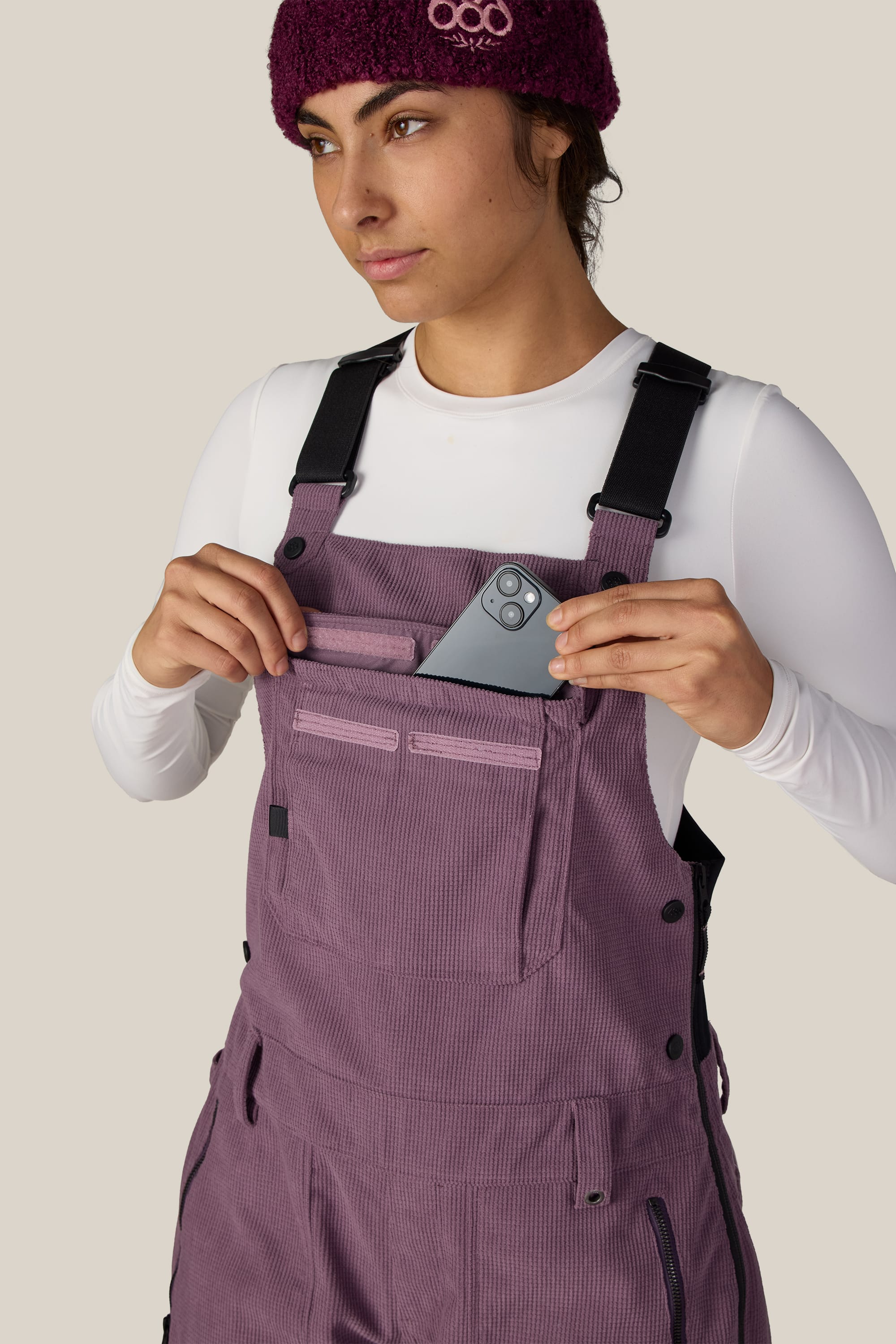 A person wearing the 686 Women’s Black Magic™ Insulated Bib by 686 places a smartphone into the front zipper pocket. They are also dressed in a white shirt, a purple knit hat, and purple overalls, looking slightly to the side.