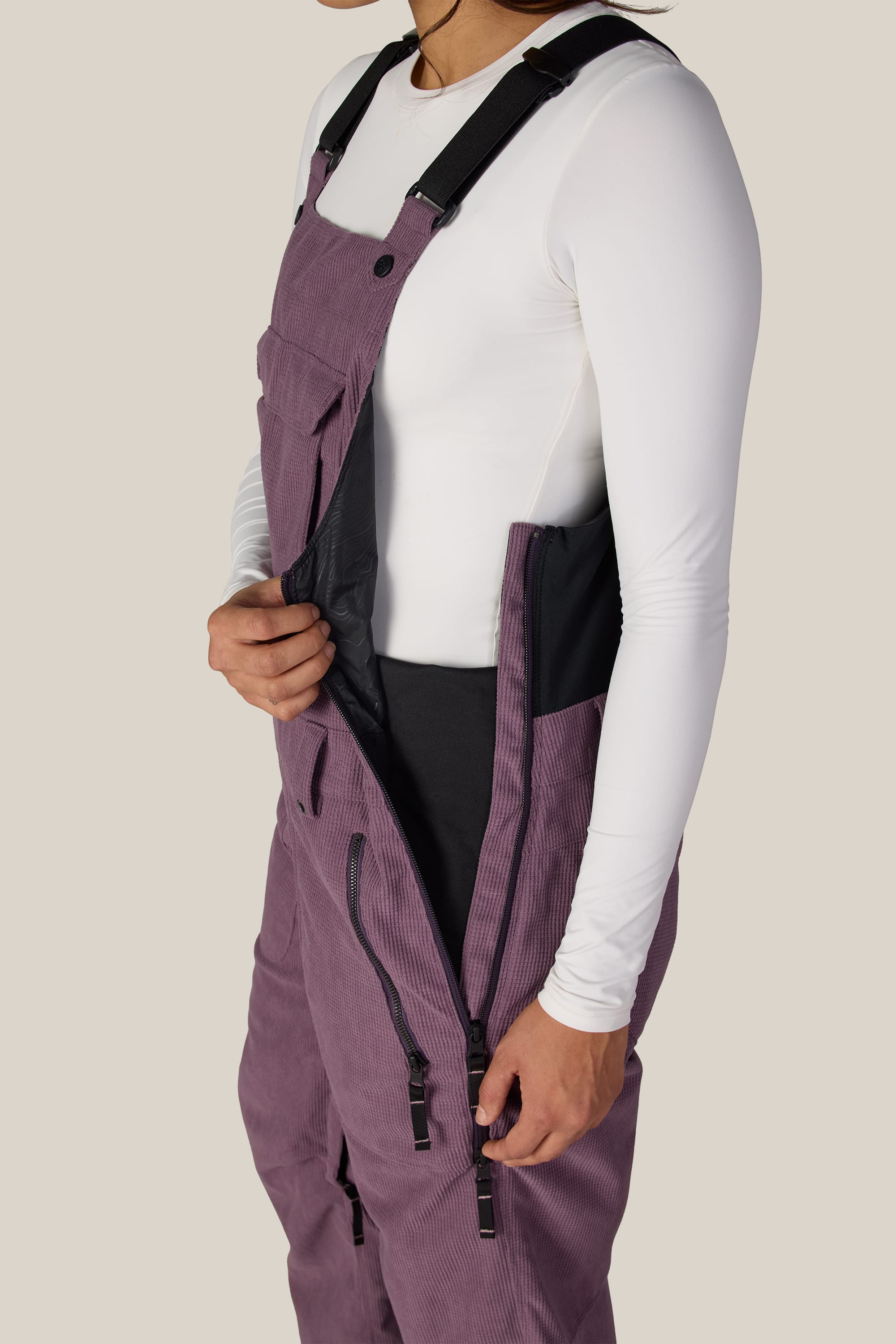 A person wearing a white long-sleeve shirt and the 686 Women’s Black Magic™ Insulated Bib is unzipping the side of the bib, revealing the black inner lining. Their head is partially out of frame.