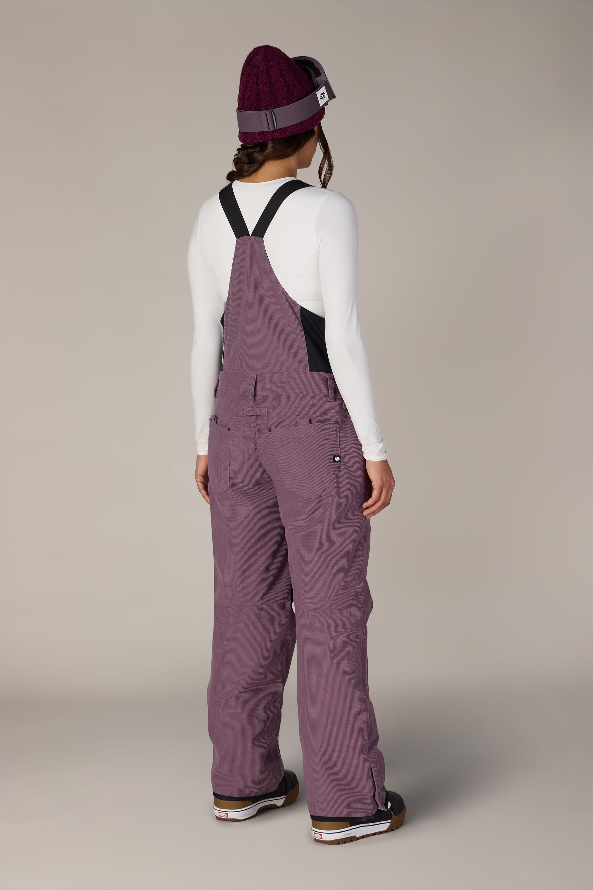 A person stands facing away, wearing the 686 Women’s Black Magic™ Insulated Bib by 686, paired with a white long-sleeve shirt, a purple knit beanie, and brown shoes against a plain light background.