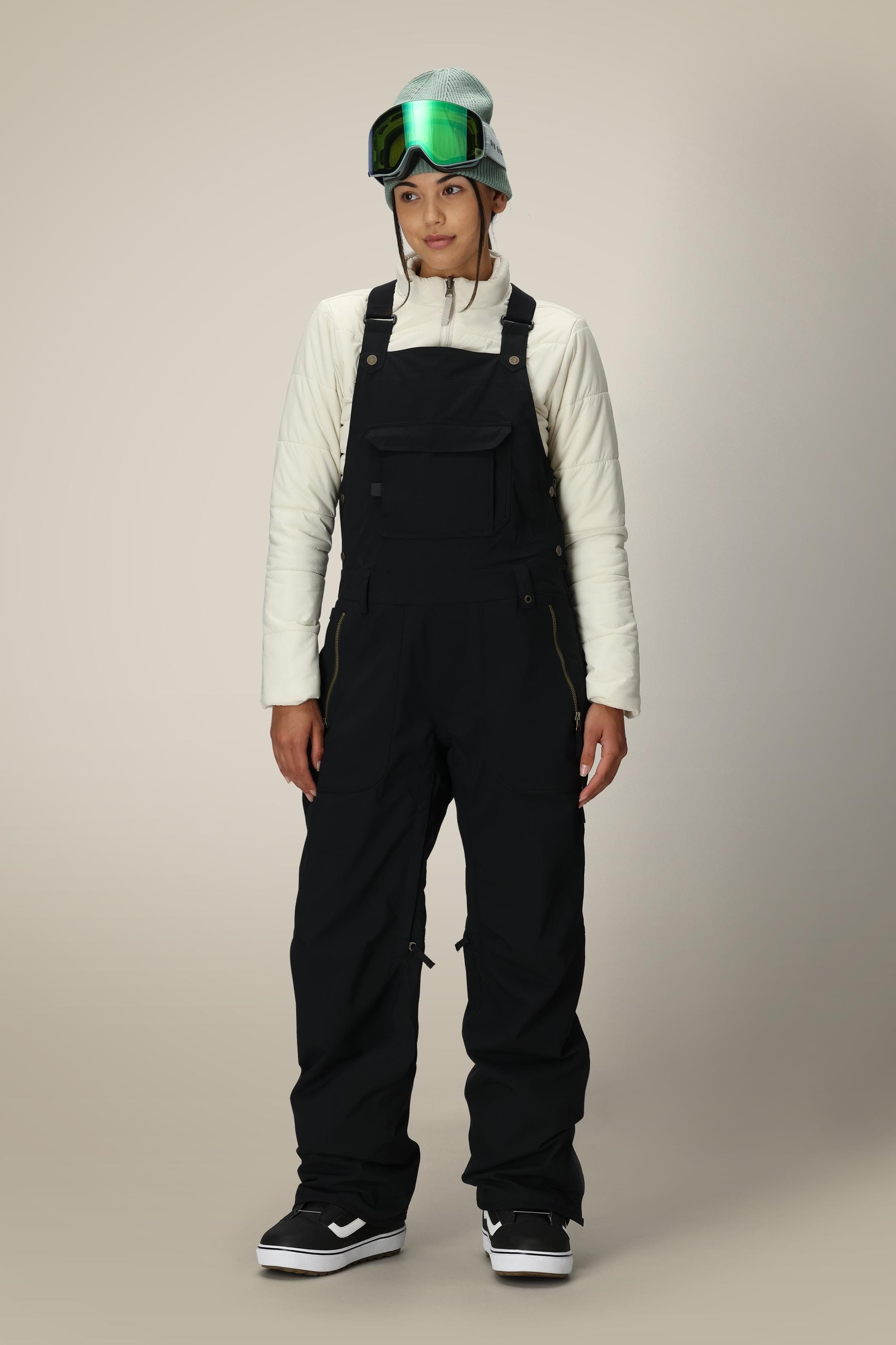 A person wearing a light long-sleeve top, 686 Women's Black Magic™ Insulated Bib, black snow boots, a blue beanie, and green ski goggles stands against a plain light background.