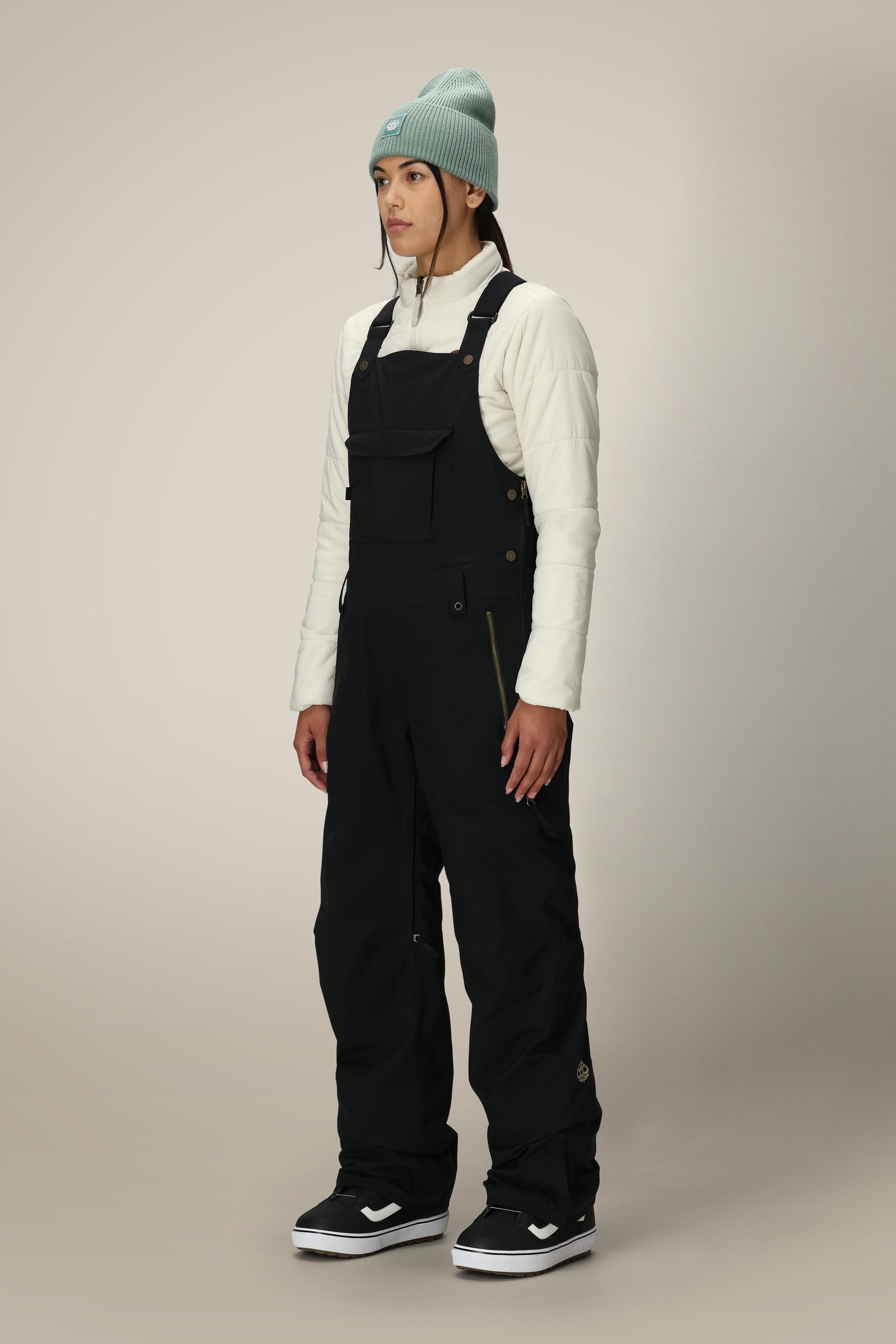 A person stands facing forward, wearing a light blue beanie, a cream quilted top, and 686 Women's Black Magic™ Insulated Bib overalls by 686 with black-and-white sneakers against a plain light background.