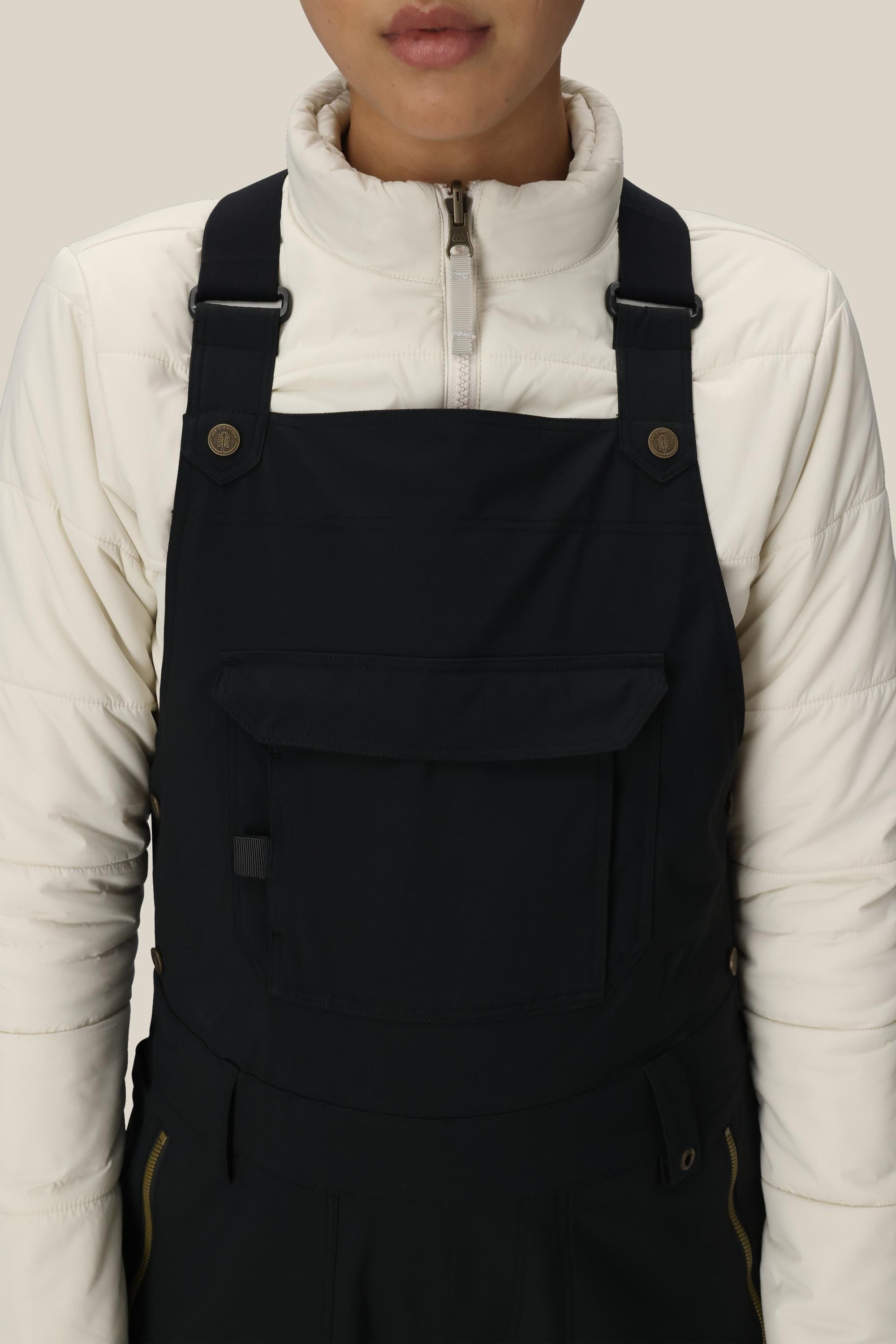 A person is shown from shoulders to waist against a plain background, wearing the 686 Women’s Black Magic™ Insulated Bib over a white zip-up jacket.