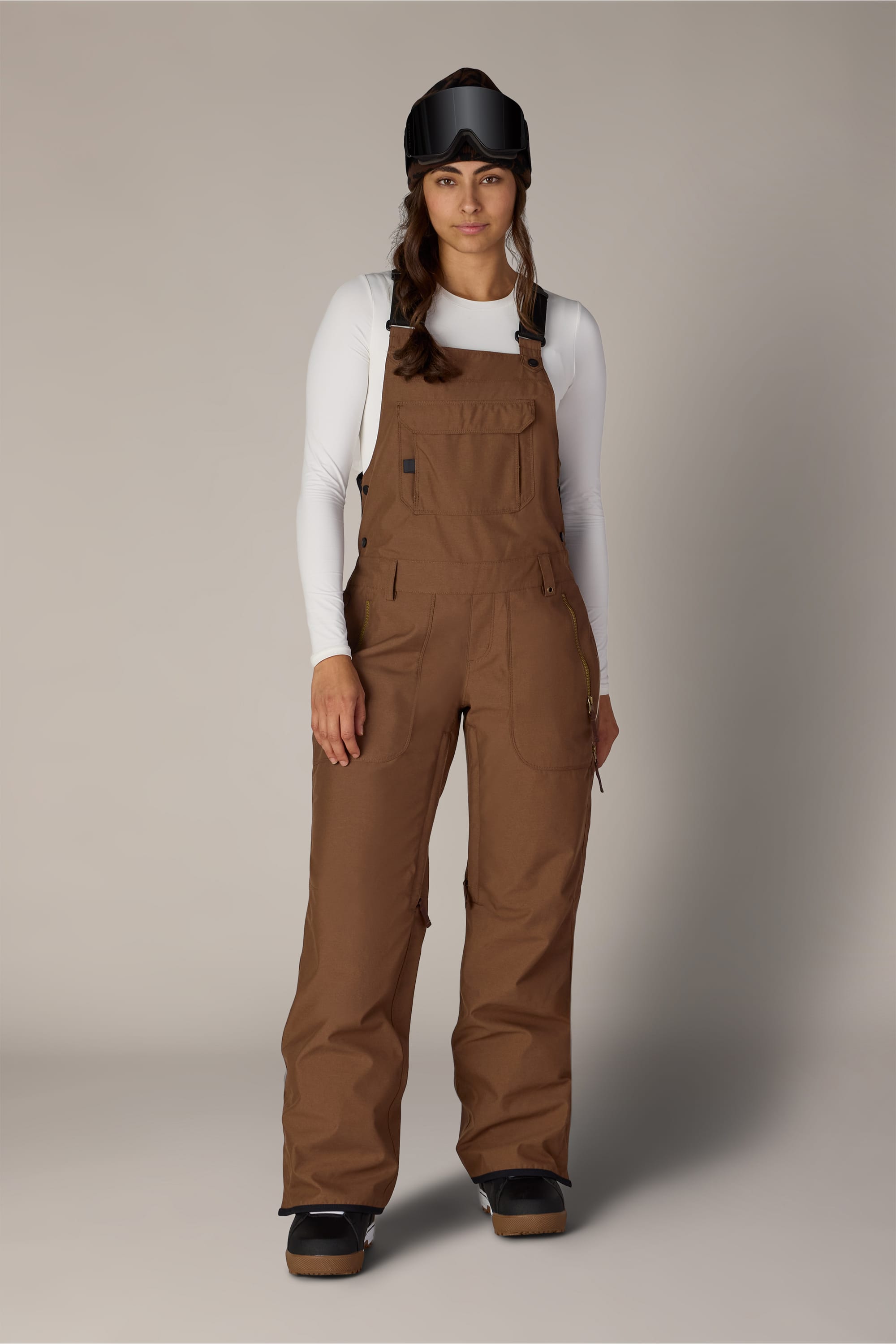 A woman stands against a plain light gray background wearing the 686 Women’s Black Magic™ Insulated Bib, paired with a white long-sleeve shirt, black snow goggles, and black shoes.