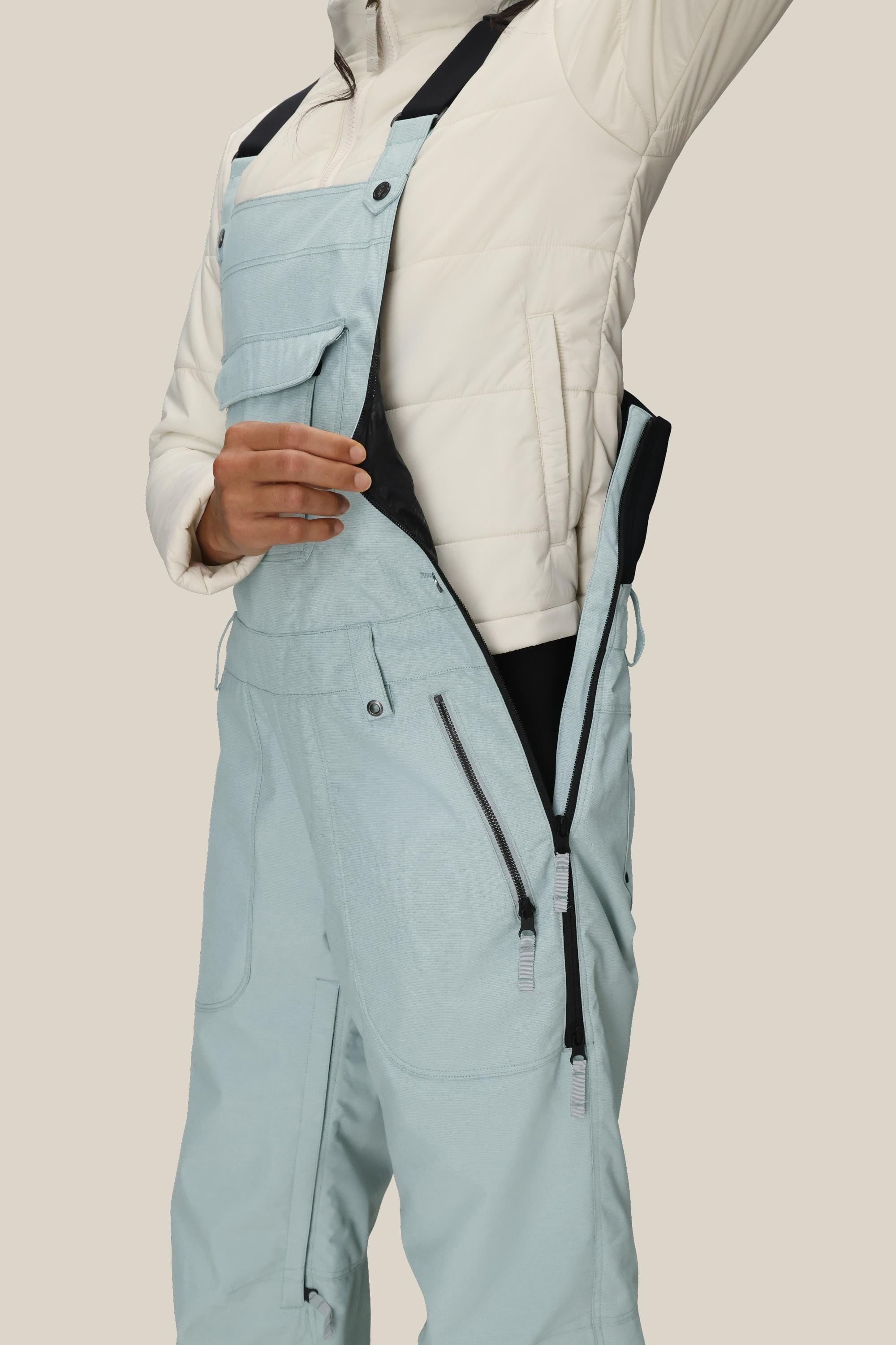 A person wearing the 686 Women's Black Magic™ Insulated Bib in light blue unzips the side to reveal a black inner lining; the image highlights the torso and upper legs against a plain background.