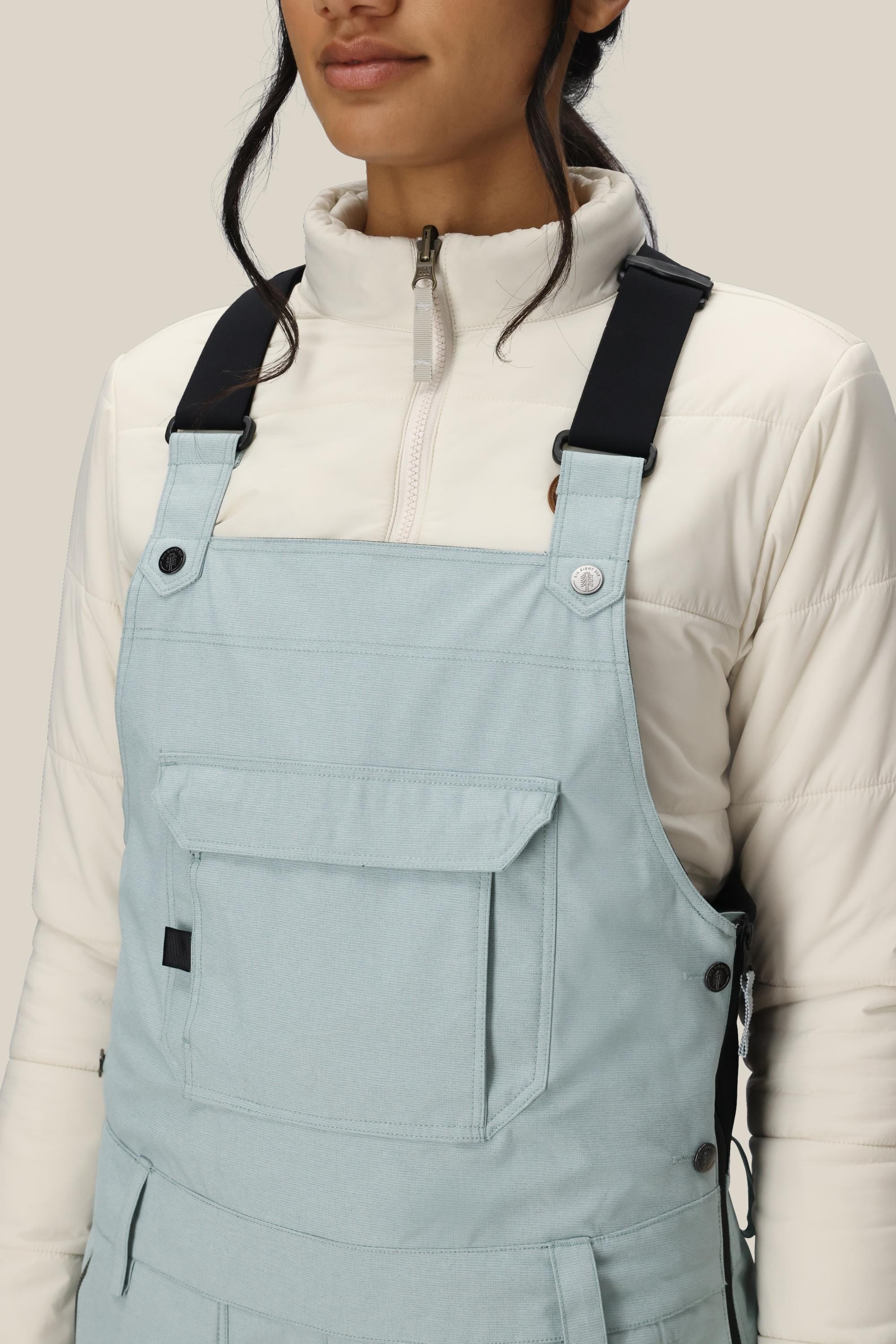 A woman wears a white zip-up jacket and 686 Women's Black Magic™ Insulated Bib overalls. The image is cropped at her shoulders, so her eyes are not visible.