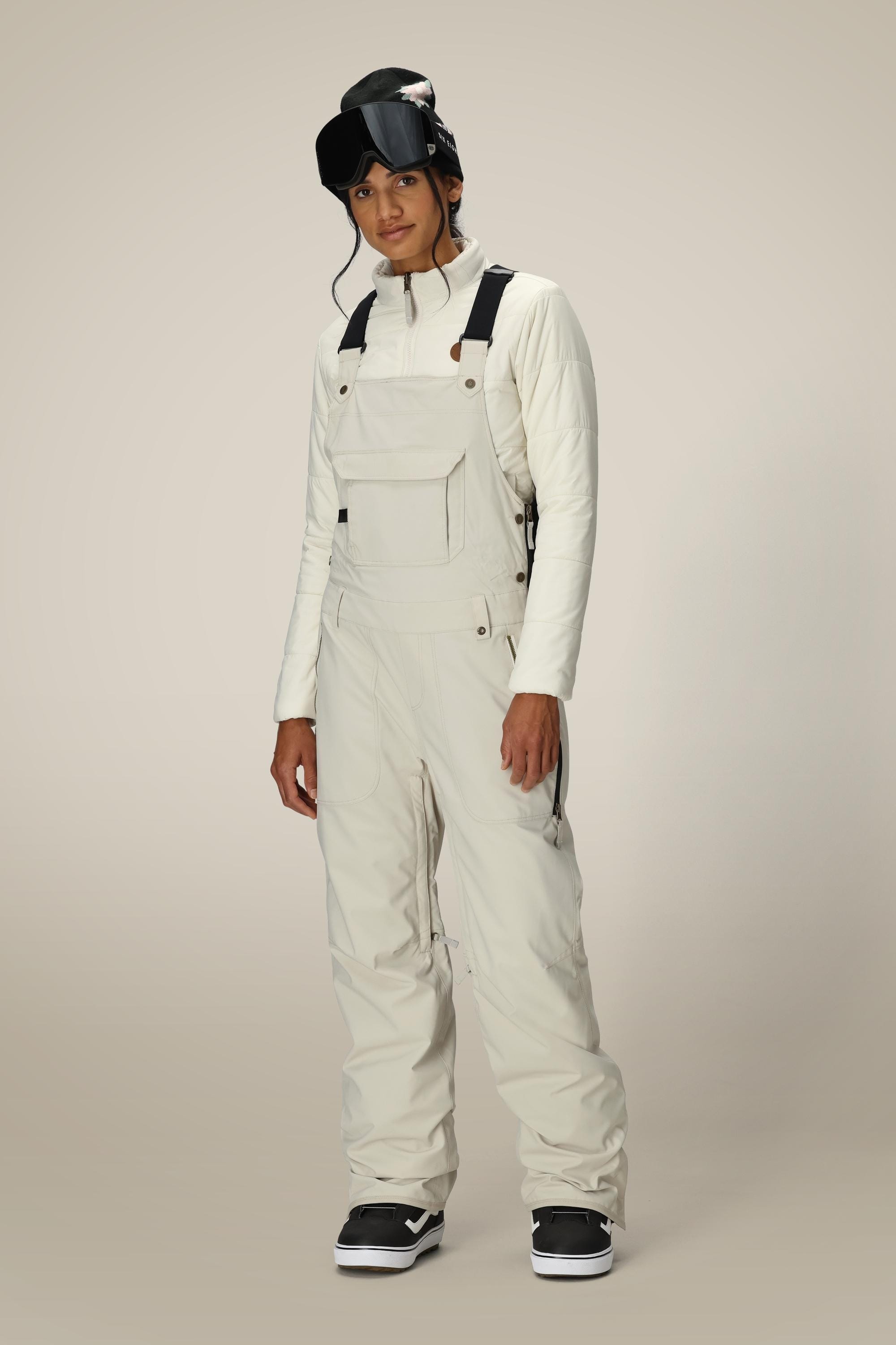 A person wearing the 686 Women’s Black Magic™ Insulated Bib by 686 stands against a plain background, dressed in an off-white winter ski outfit with matching jacket, gloves, and black ski goggles resting on their head.