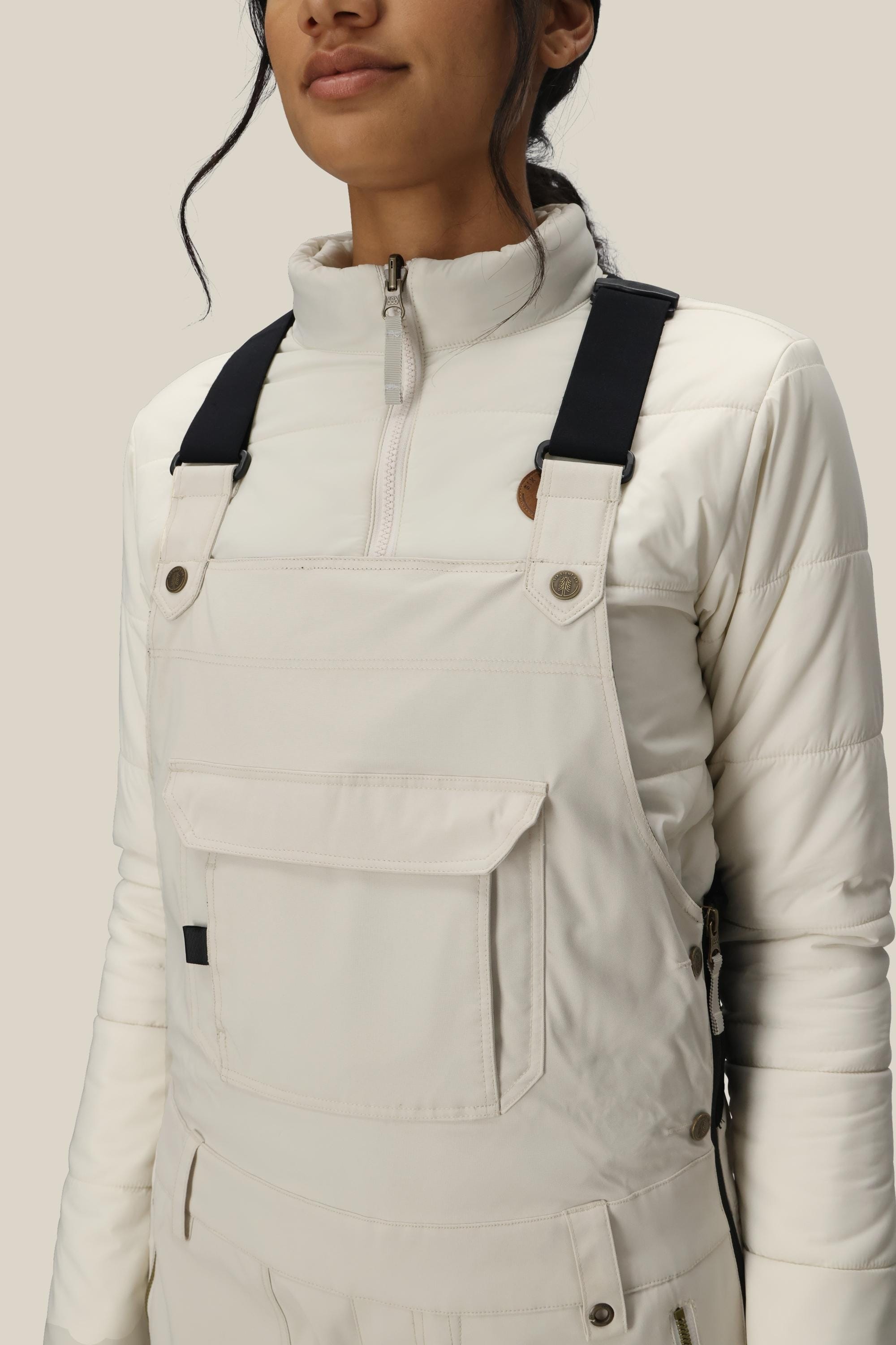 A woman wearing the 686 Women’s Black Magic™ Insulated Bib, featuring black straps, paired with a white quilted jacket and standing against a plain light background. Only her lower face and torso are visible.