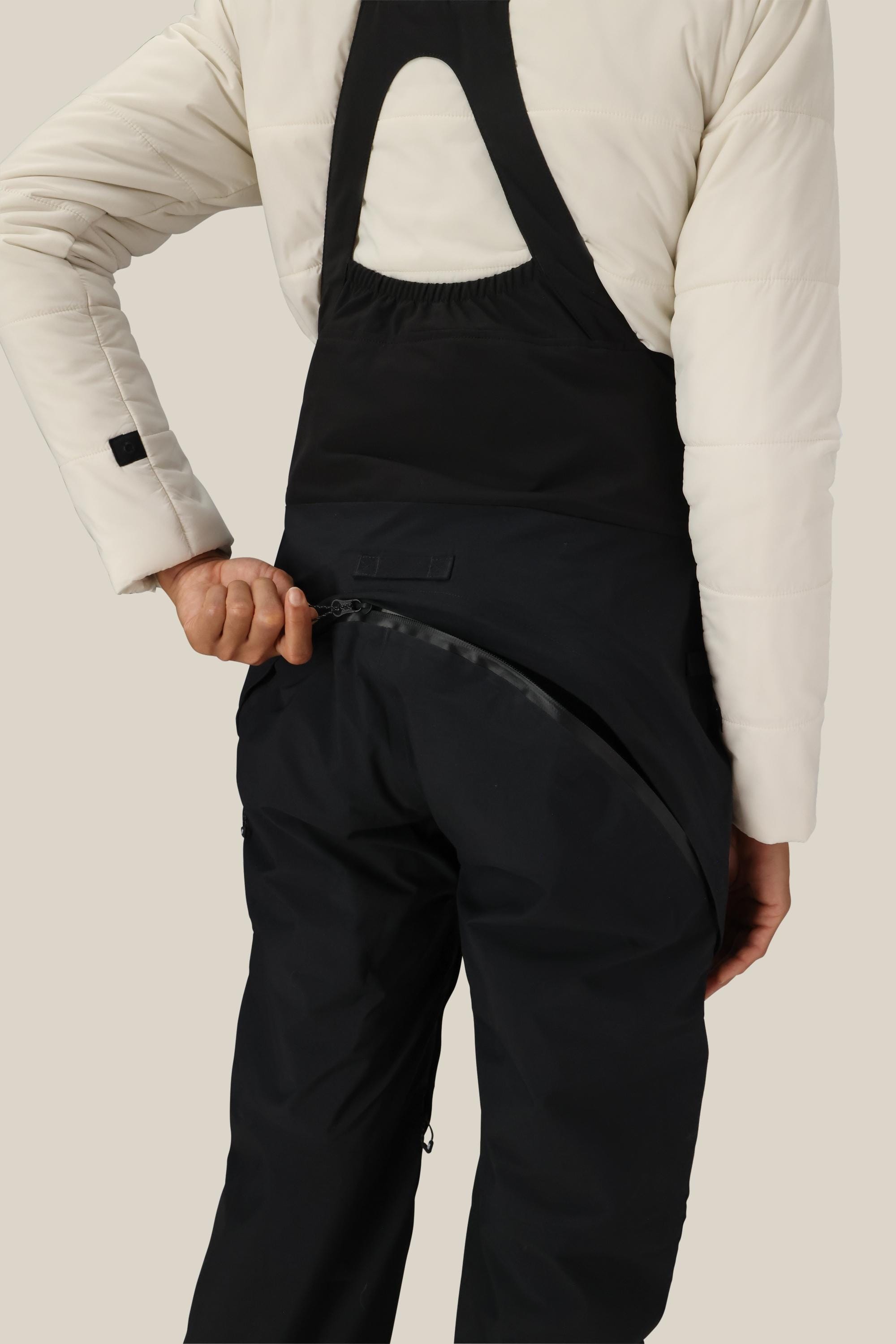 A person wearing the 686 Women's GORE-TEX Dispatch™ Shell Bib in black and a white jacket unzips the large diagonal back zipper, showcasing the design and functionality of this 686 garment.