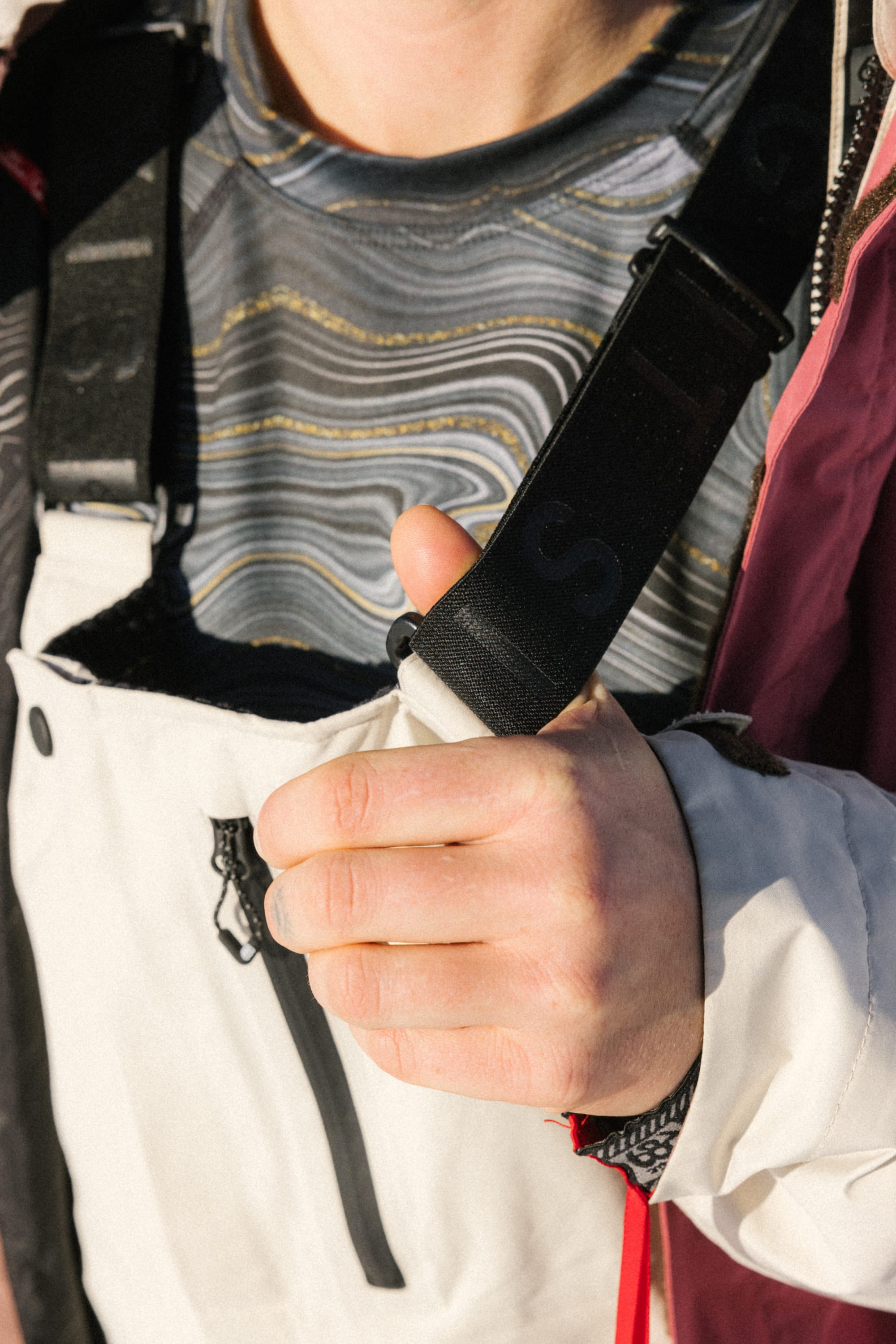 Wearing a patterned shirt and winter jacket, a person adjusts the strap on their 686 Women's GORE-TEX Dispatch™ Shell Bib, sunlight highlighting their hand and upper body.
